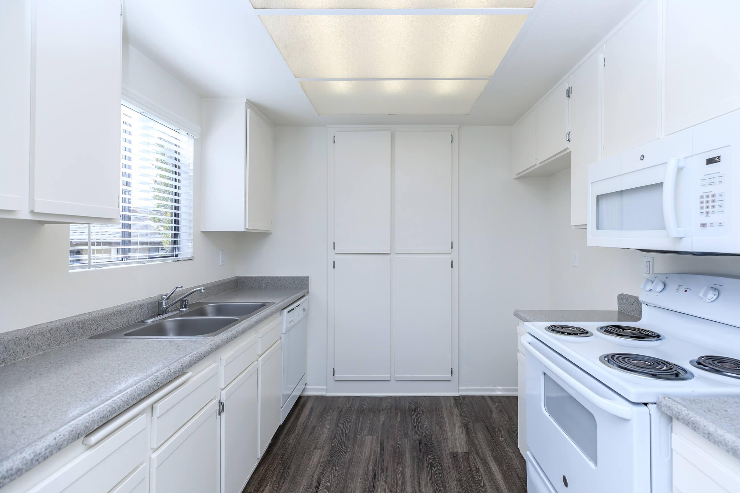 Bright, modern kitchen featuring white cabinetry, a double sink, and gray countertops. Equipped with a stove, microwave, and a wall cabinet for storage. The large window allows natural light to flood the space, enhancing the clean and spacious feel of the room. The flooring is dark wood laminate.