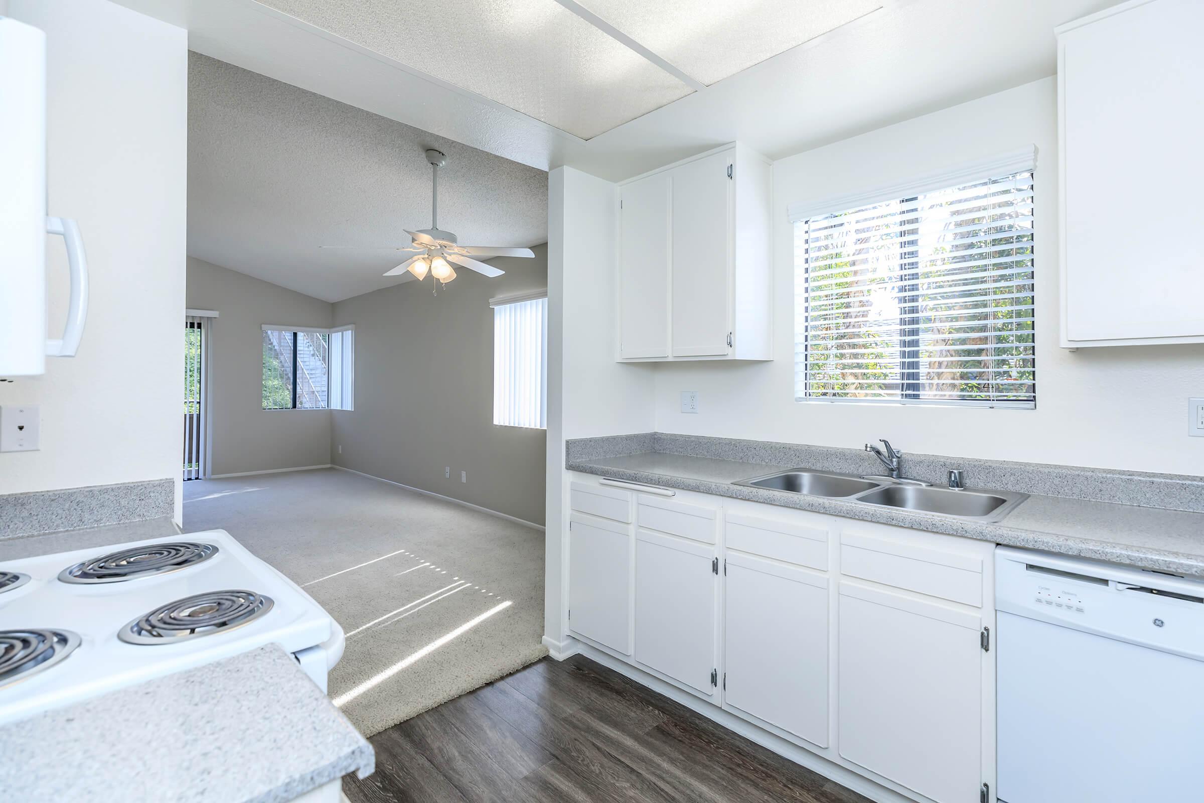 Bright kitchen with white cabinetry and a two-basin sink, featuring a stove with coil burners. Natural light streams through a window above the sink. In the background, a spacious living area with neutral carpeting and a ceiling fan is visible, along with another window that opens to the outdoors.