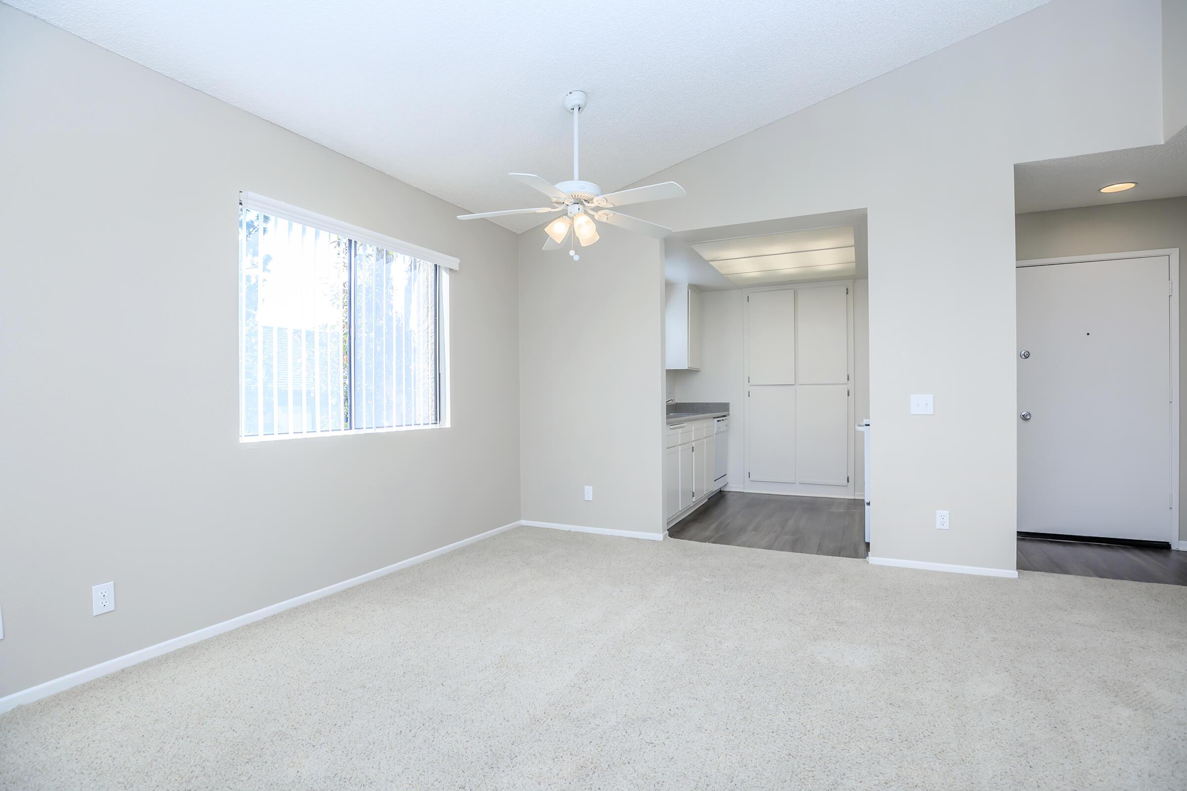 A spacious, light-filled living area with beige carpet and neutral-colored walls. There is a ceiling fan and a large window allowing natural light. A doorway leads to a kitchen area with white cabinetry, and an entryway with a door is visible on the right. The room offers a clean, contemporary aesthetic.