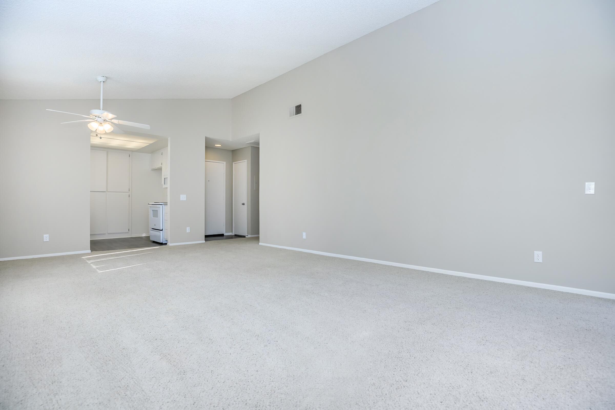 Spacious, empty living room with light gray walls and beige carpet. A ceiling fan is installed, and a doorway leads to a kitchen area with white appliances. The room has a minimalist design, with no furniture, creating an open and airy atmosphere.