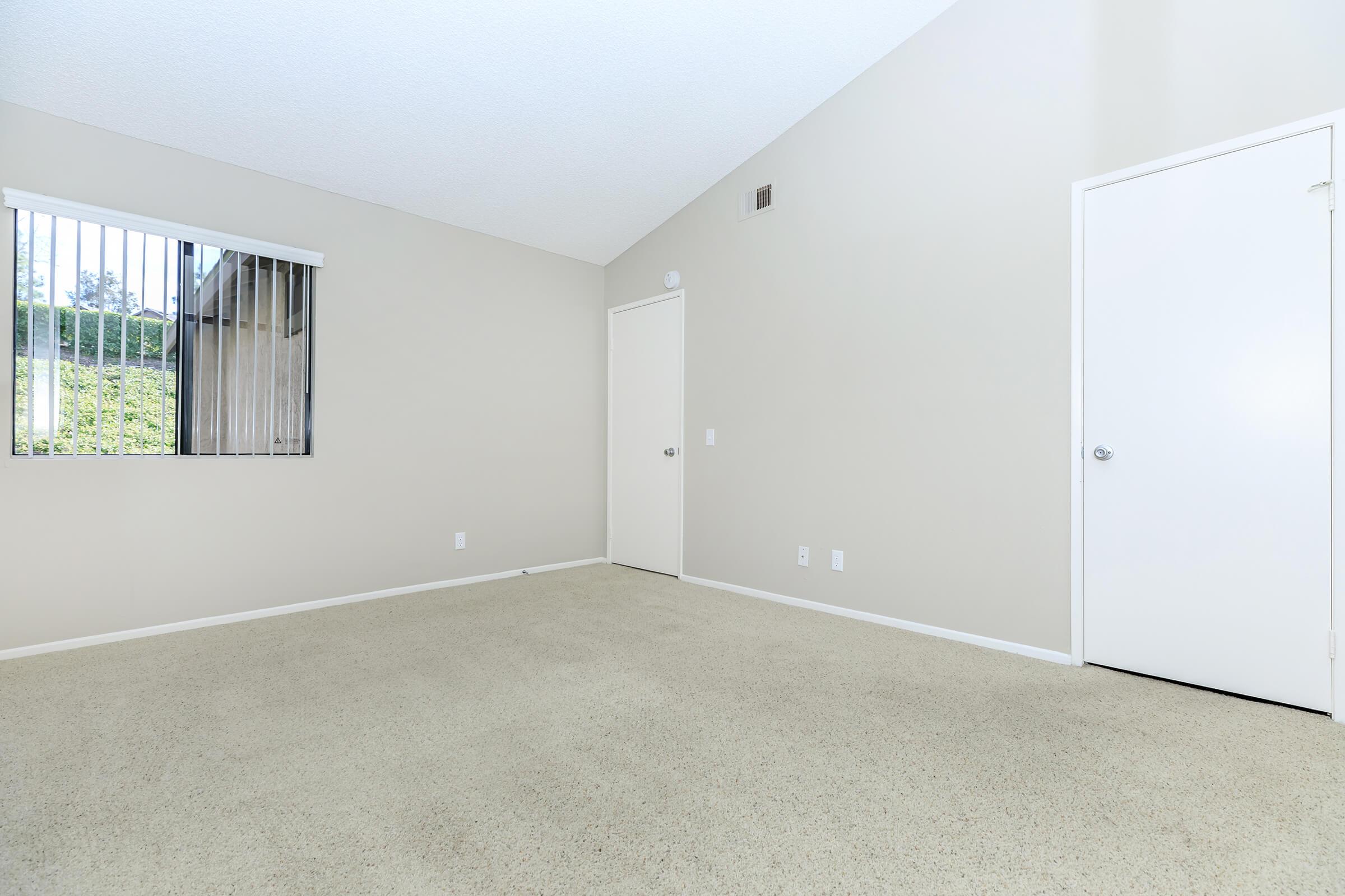 A bare room with light beige walls and a carpeted floor. A window with horizontal blinds is on the left, and a closed door is on the right. The ceiling is sloped, and the overall lighting is bright and neutral, creating a spacious and inviting atmosphere.