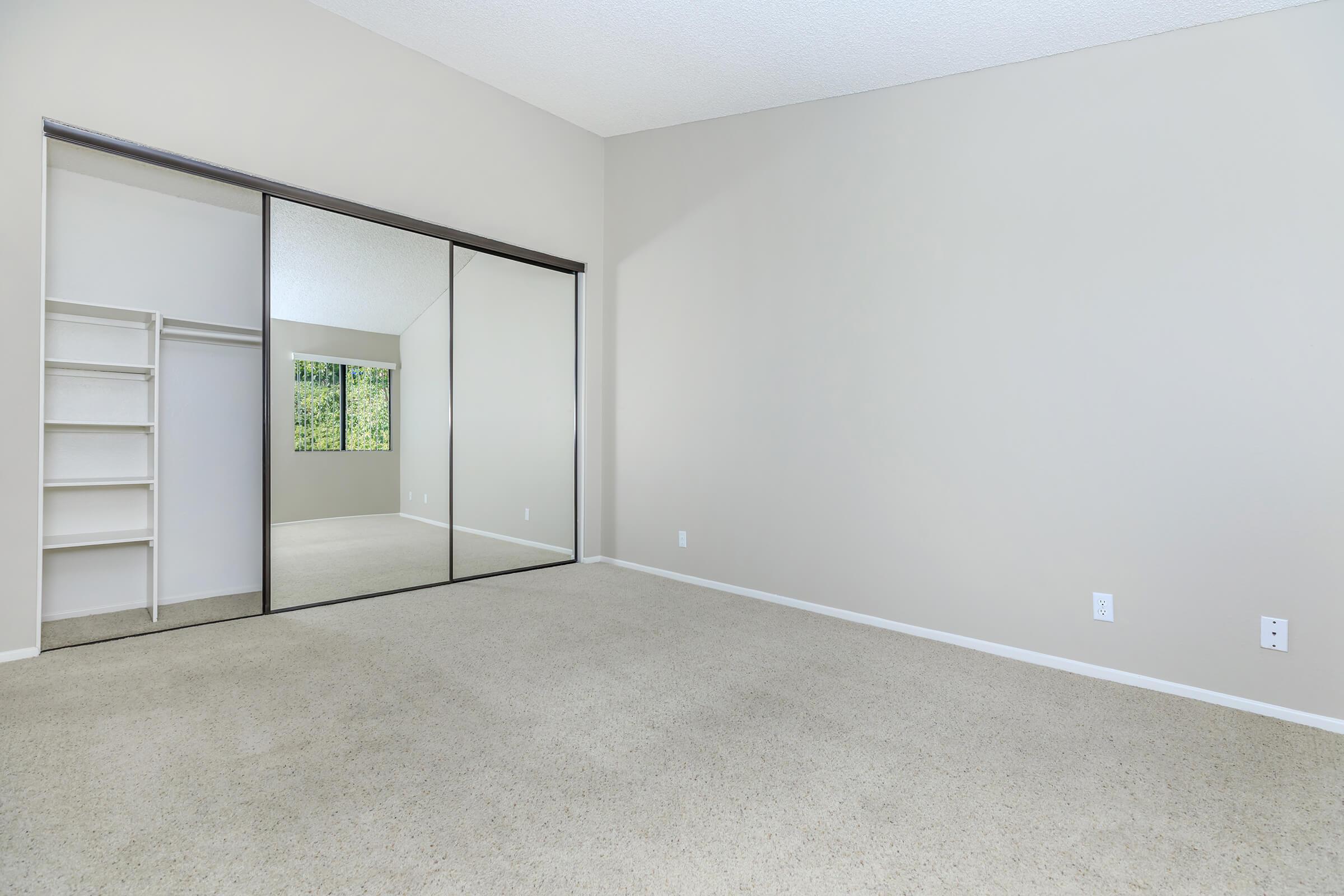A spacious, empty room with light-colored walls and carpet. There is a large mirrored closet on one side and a window allowing natural light. The room has a neutral aesthetic, with ample space for furniture and decoration. The overall atmosphere is bright and inviting.