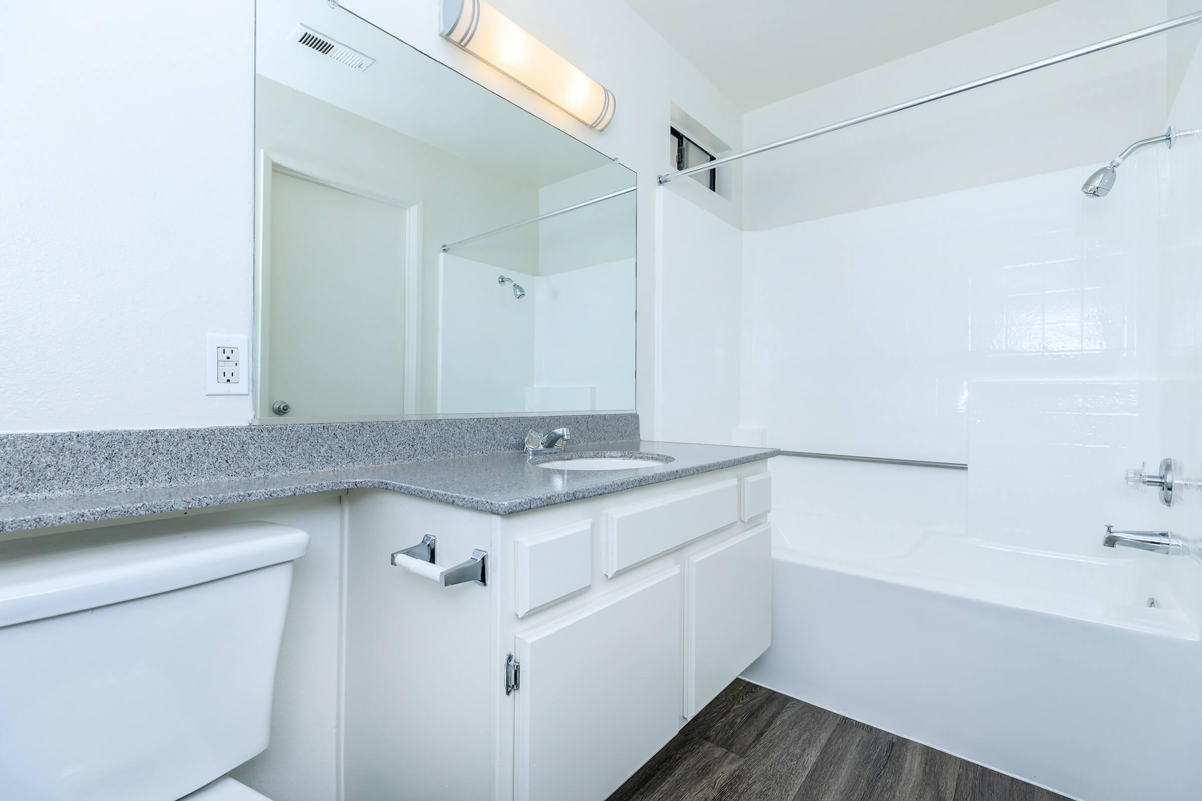 A clean and modern bathroom featuring a white bathtub and shower, a white sink with a granite countertop, and a mirror above the sink. The floor is a dark wood-like material, and there is a light fixture on the wall. The space is bright and well-lit, with simple and contemporary design elements.