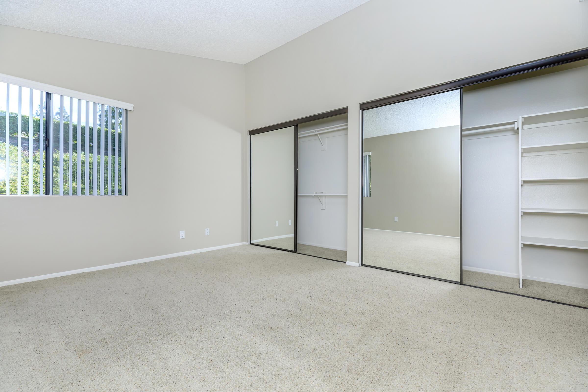 A bright, empty room with beige walls and carpet, featuring a window with vertical blinds. There are two mirrored sliding closet doors on one wall and an open shelf unit beside them, creating a spacious and airy feel. The natural light enhances the room's openness.