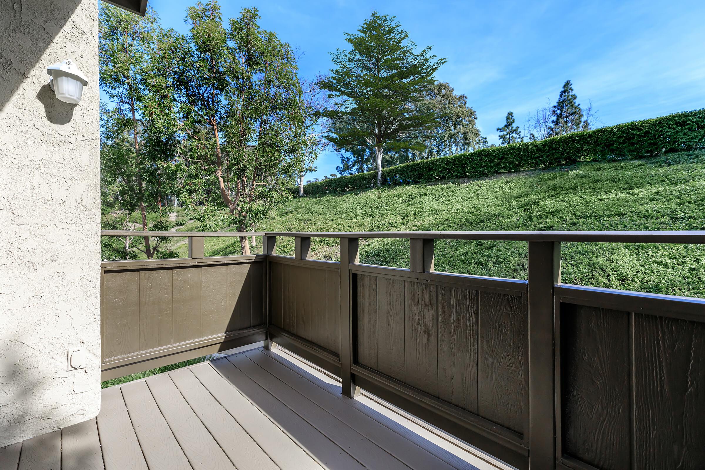 A view from a balcony overlooking a lush green hillside, with trees and shrubs in the background. The balcony features brown wooden railing and flooring, providing a peaceful outdoor space. Bright blue sky can be seen above, indicating a clear day.