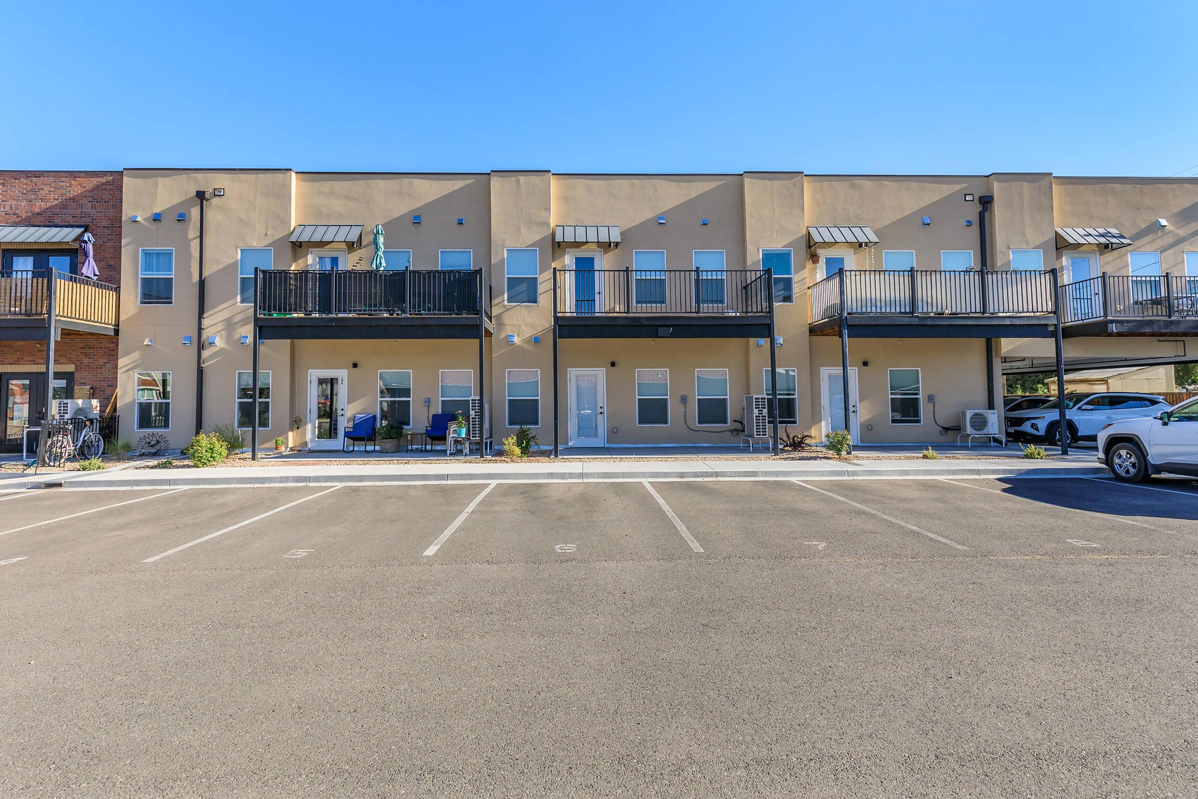A row of modern apartment buildings with balconies, featuring a light-colored exterior. The ground level has several doors leading to individual units. In front, there are empty parking spaces and some bicycles. The scene is set under a clear blue sky.