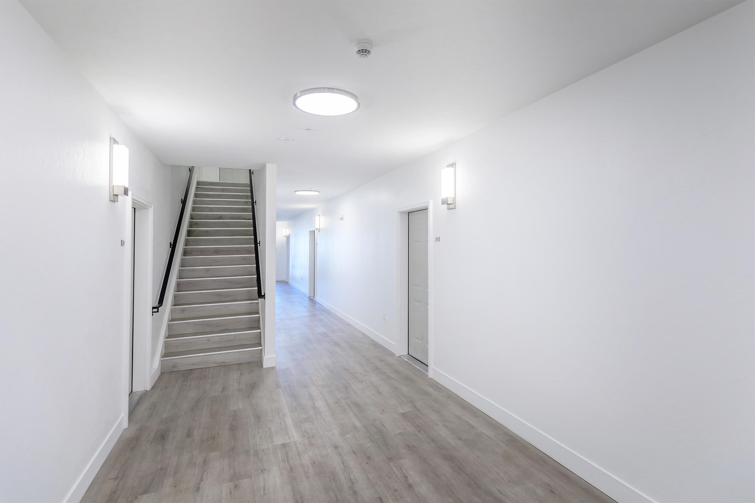 A well-lit corridor with a staircase on the left, featuring white walls and wooden flooring. There are doors on both sides of the hallway, and modern light fixtures illuminate the space, creating a clean and minimalist atmosphere.