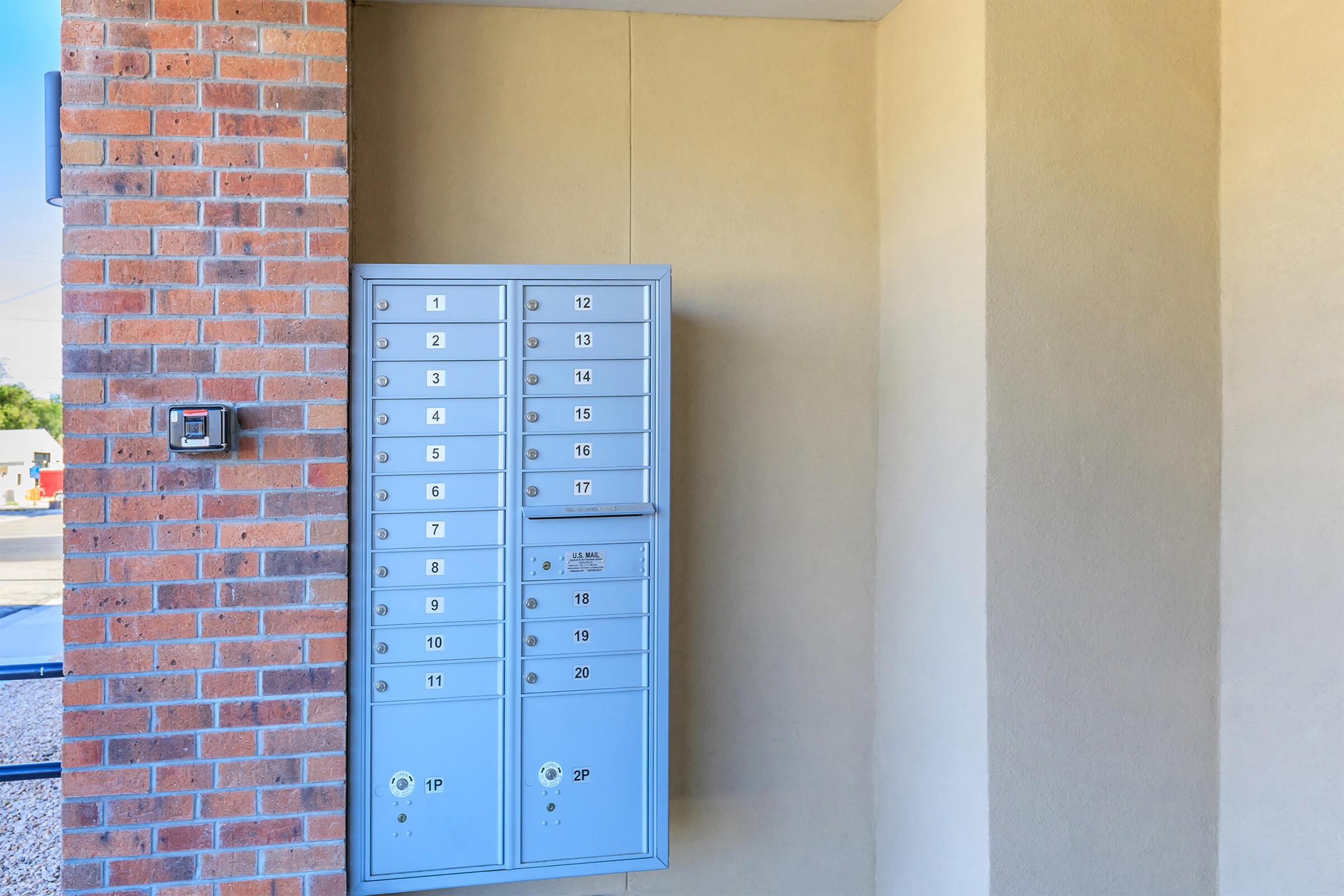 A cluster of outdoor mailboxes mounted on a wall, featuring multiple numbered slots for mail delivery. The wall is a light beige color, and the adjacent section is made of red brick. A small keypad is also visible near the mailboxes.