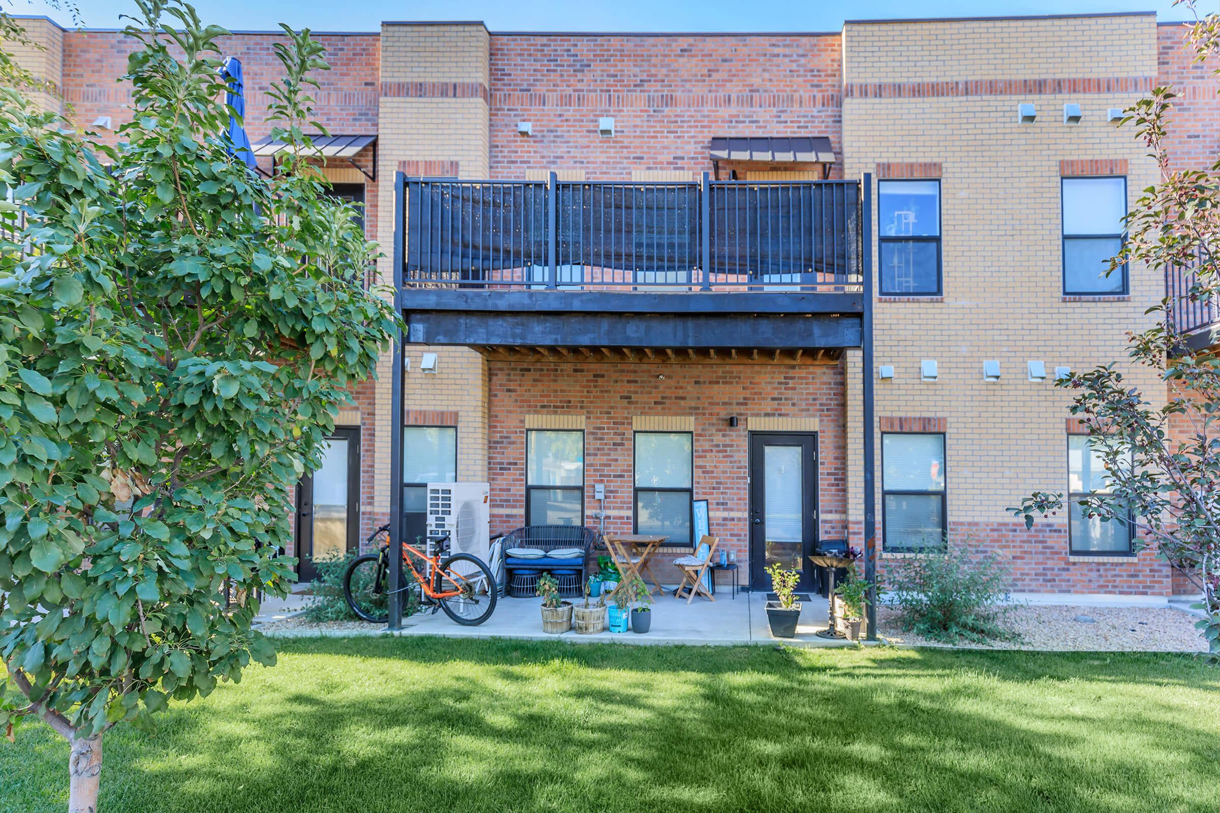 A modern multi-story apartment building features a ground-level patio with a bicycle, a small table, and decorative plants. The structure has brick and wood accents, large windows, and a balcony above. Lush green grass and trees surround the outdoor space, creating a pleasant residential environment.