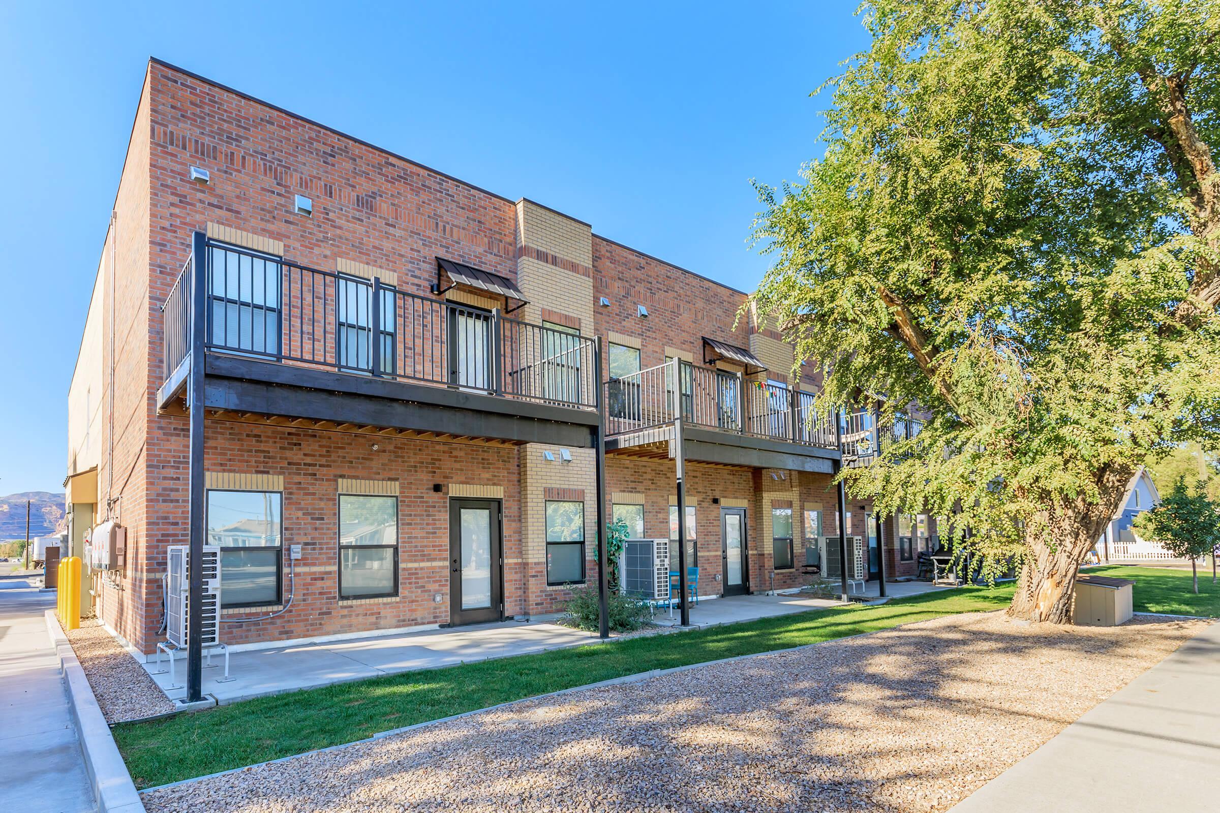 Modern brick building with multiple apartments featuring balconies and large windows. A green lawn and trees are in the foreground, while a clear blue sky provides a bright backdrop. The design reflects contemporary architecture with an inviting exterior.