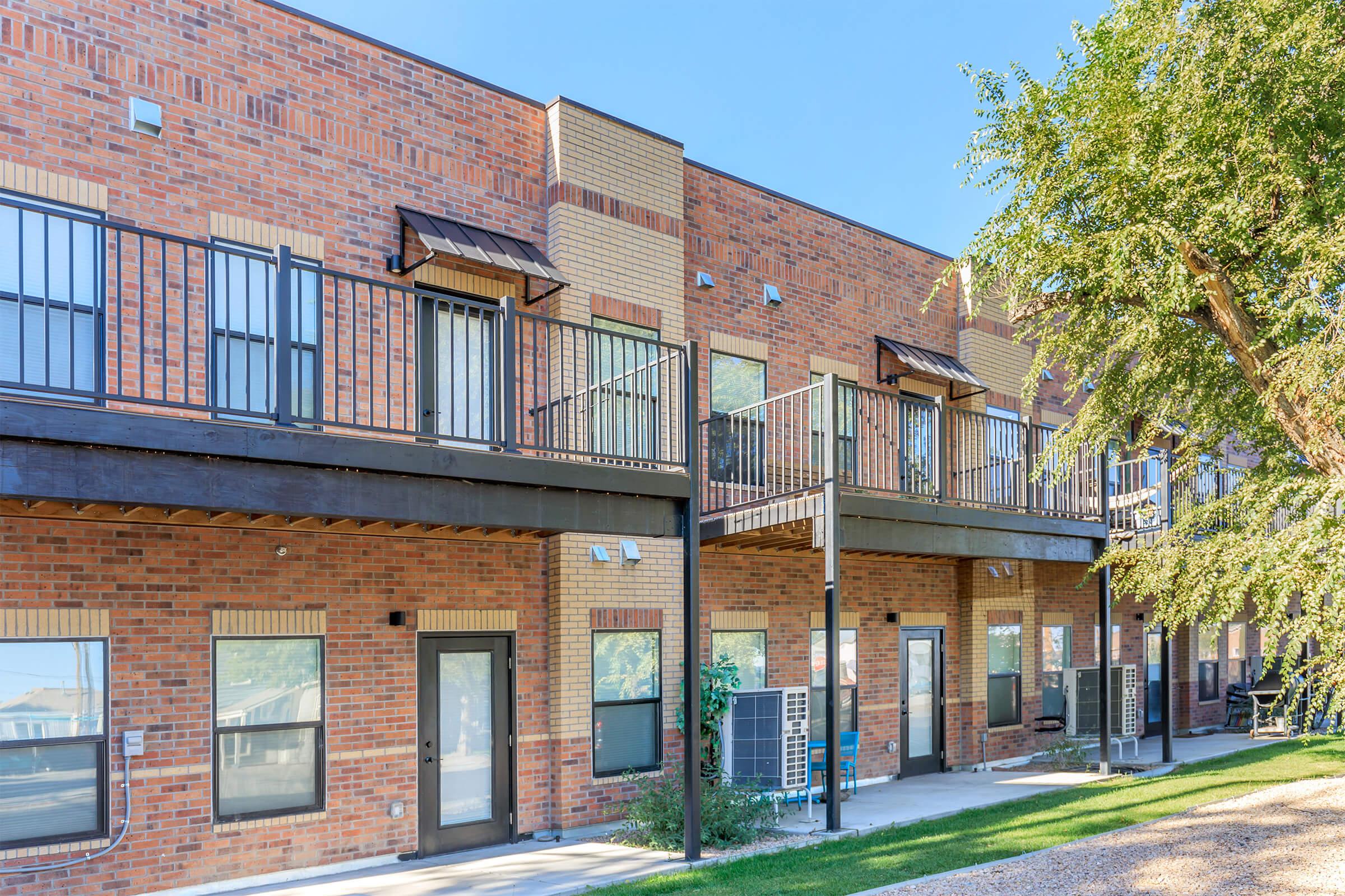 A row of modern townhomes featuring brick exteriors and balconies. Each unit has large windows and a private entrance, with green grass and decorative trees in the foreground. Air conditioning units are visible below the balconies, enhancing the homey appearance of the buildings. Clear blue sky above.