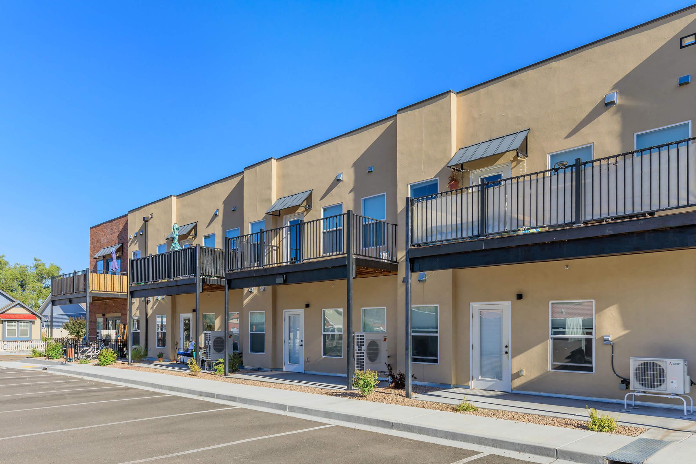 A row of modern, two-story residential buildings featuring balconies and air conditioning units. The facade is tan with blue accents, and the area is landscaped with gravel and minimal greenery. The parking lot in front is well-defined with individual parking spaces. Clear blue sky overhead.
