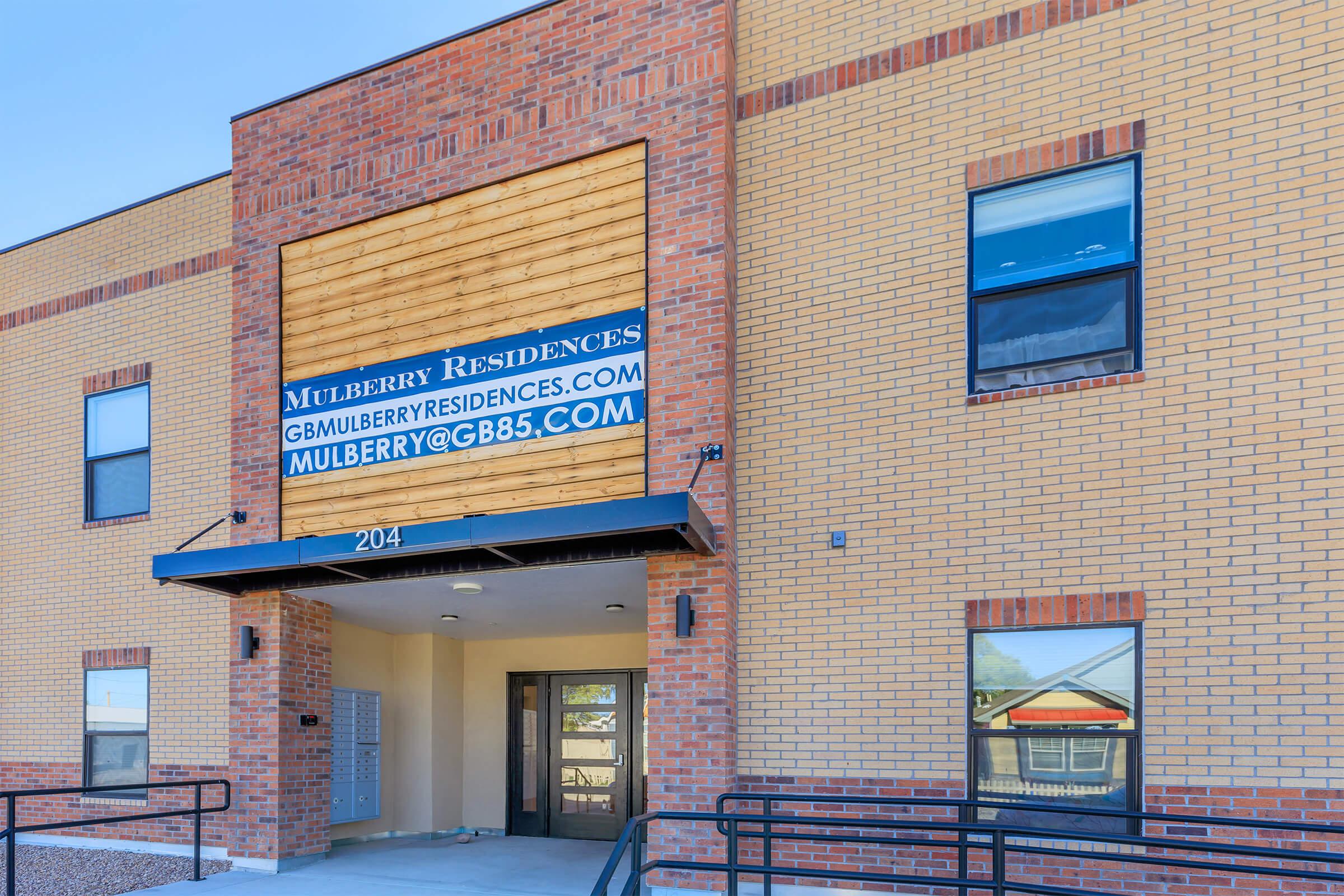 Exterior view of the Mulberry Residences building featuring a modern design with brick walls and large windows. The entrance includes a sign displaying the website and email contact for inquiries. A ramp leads to the entrance, ensuring accessibility. Bright daylight enhances the welcoming appearance of the building.