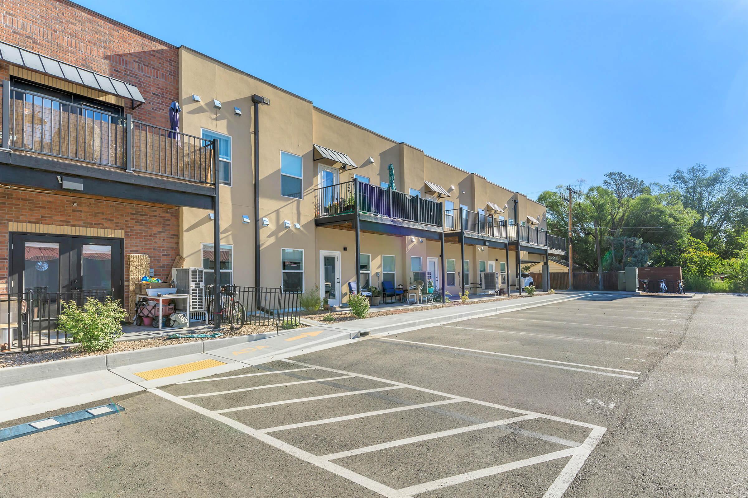 View of a multi-story building with a mix of modern architectural designs. The building features balconies and several entrance doors, surrounded by a paved parking area. Sunlight illuminates the scene, enhancing the greenery in the background and the clear blue sky above.