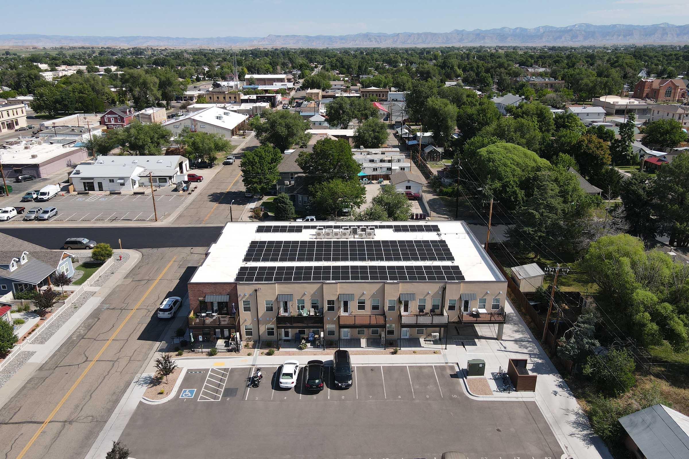 Aerial view of a small-town commercial building with solar panels on the roof. Surrounding the building are trees and other buildings, with a parking lot in the foreground. The landscape features distant mountains and a clear sky, indicating a rural or suburban area.
