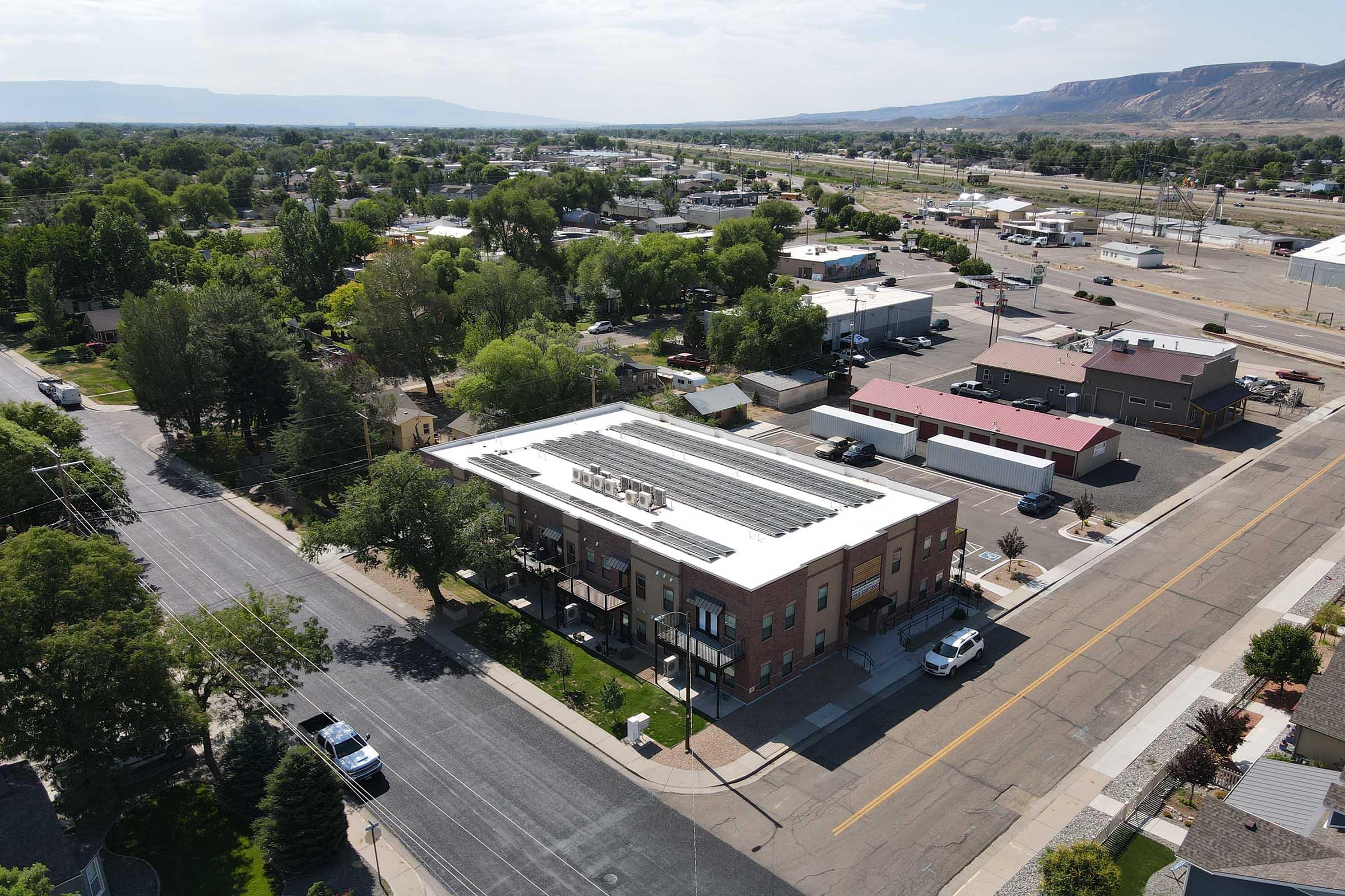 Aerial view of a small town, featuring a modern building with solar panels on the roof, surrounded by trees and other commercial structures. Streets are visible, with vehicles parked and driving by. Mountains can be seen in the background, indicating a scenic location.