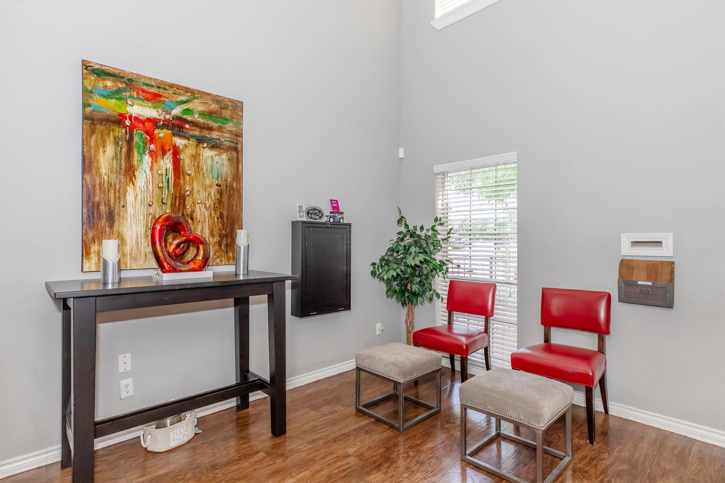 A modern living space featuring a gray wall, a colorful abstract painting, a black console table, two red chairs, and two gray stools. There is a small green plant by the window and a wall-mounted mailbox nearby, creating a welcoming atmosphere.