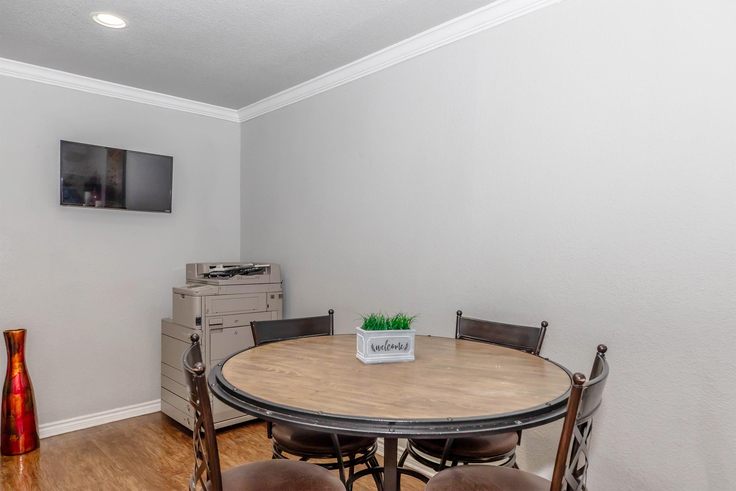A round wooden dining table surrounded by four brown chairs, with a small plant in a rectangular pot on the table. In the background, there's a TV mounted on the wall and a filing cabinet. A decorative vase in a vibrant color is positioned on the left side of the image. The walls are painted light grey.