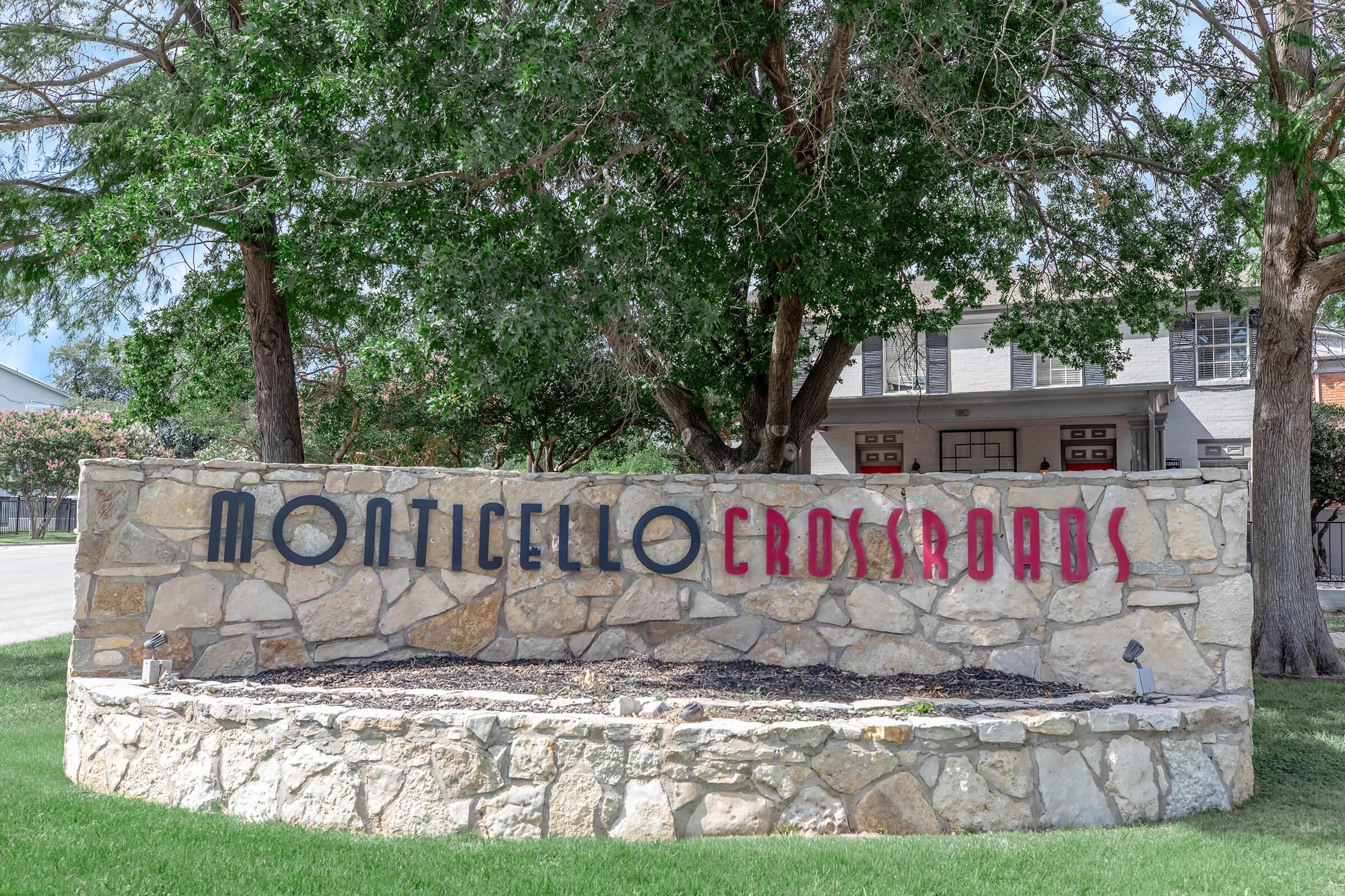 Sign for Monticello Crossroads, made of stone, displaying the words "MONTICELLO" in large, bold letters and "CROSSROADS" in a smaller, contrasting font. The setting includes green grass and trees in the background, with a clear blue sky.