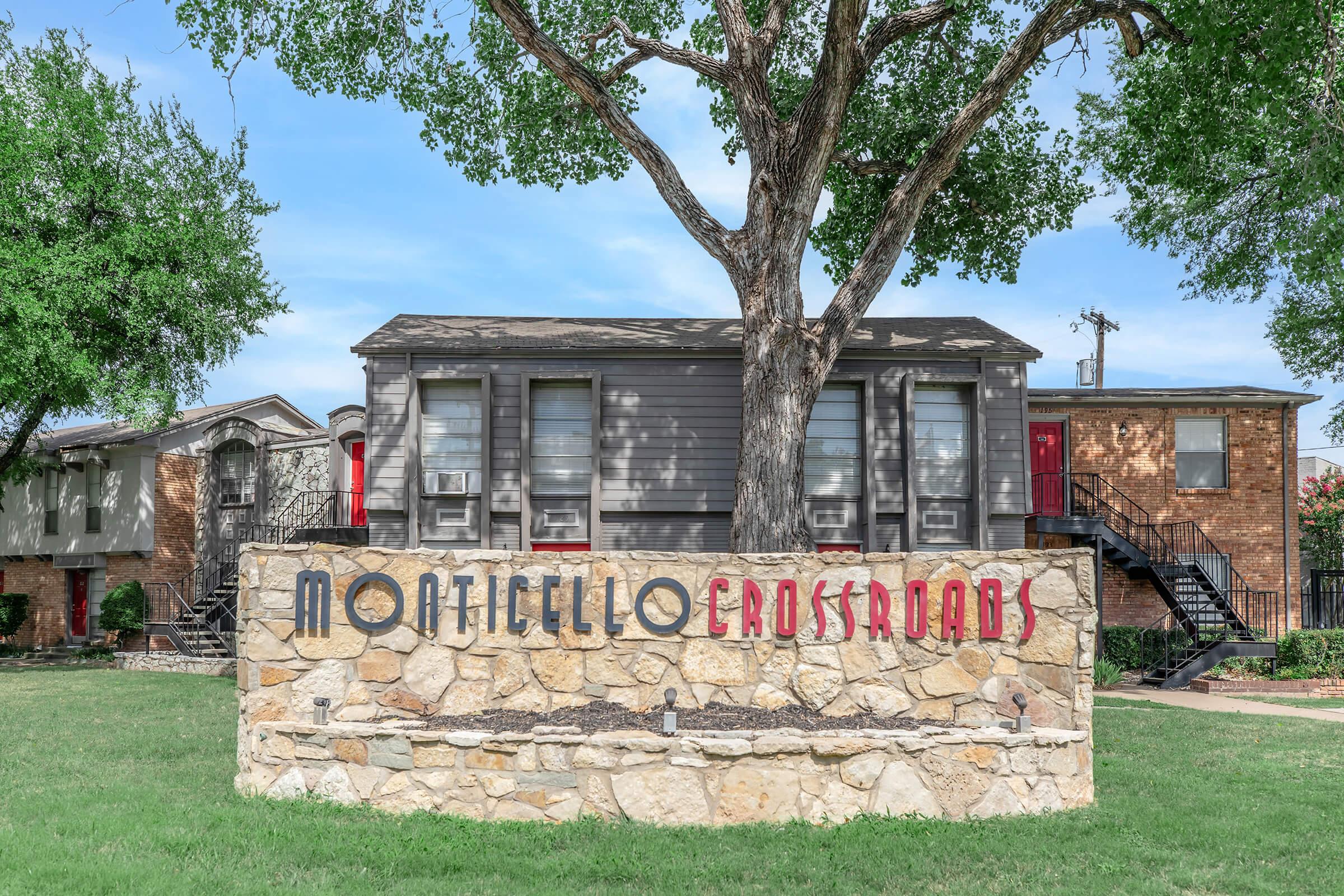 A stone sign reading "Monticello Crossroads" in red letters, located in front of a two-story building with air conditioning units and a tree nearby. The setting features a green lawn and neighboring buildings in the background.