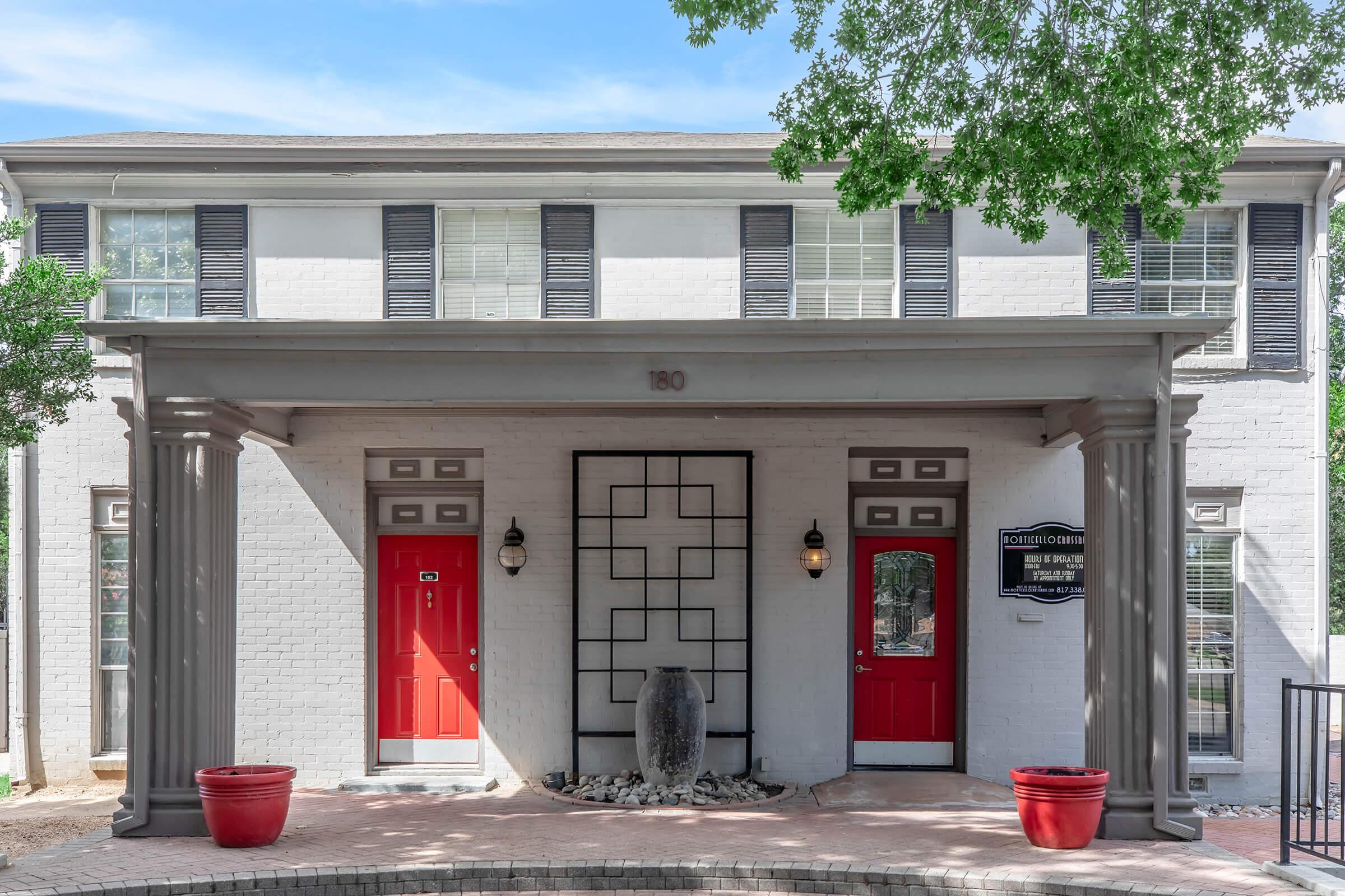 A two-story building with a white brick facade, featuring two red doors on either side of a central structure adorned with decorative elements. A pathway with a circular planter at the front leads to the entrance, and green trees provide shade around the area.
