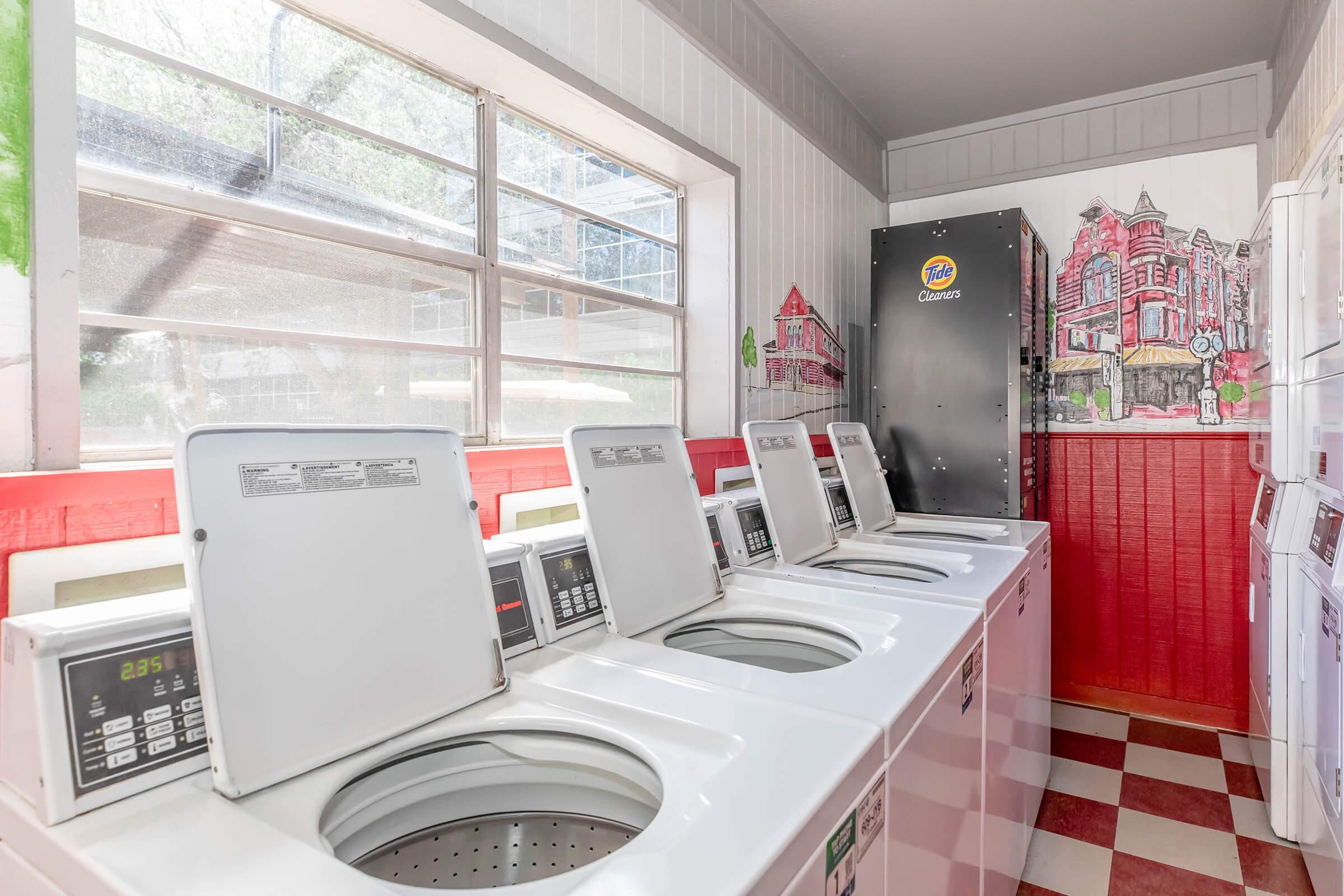 A bright laundry room featuring several white washing machines and a dryer, with a large window allowing natural light. The walls are adorned with a colorful mural depicting a cityscape, and the floor is covered in red and white checkered tiles.