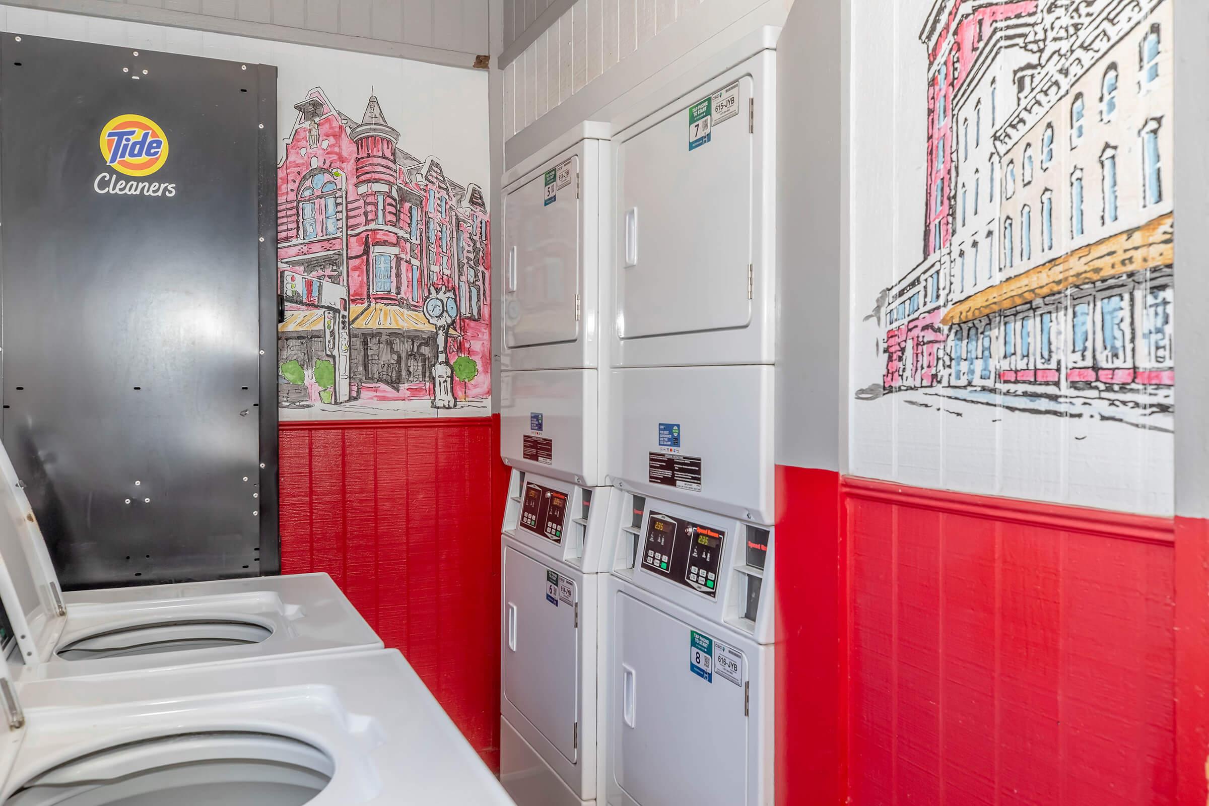 Interior of a laundry facility featuring modern washing machines and dryers. The walls are adorned with colorful illustrations of buildings, adding a lively touch to the space. A Tide Cleaners logo is visible in the corner, emphasizing the laundry service theme.