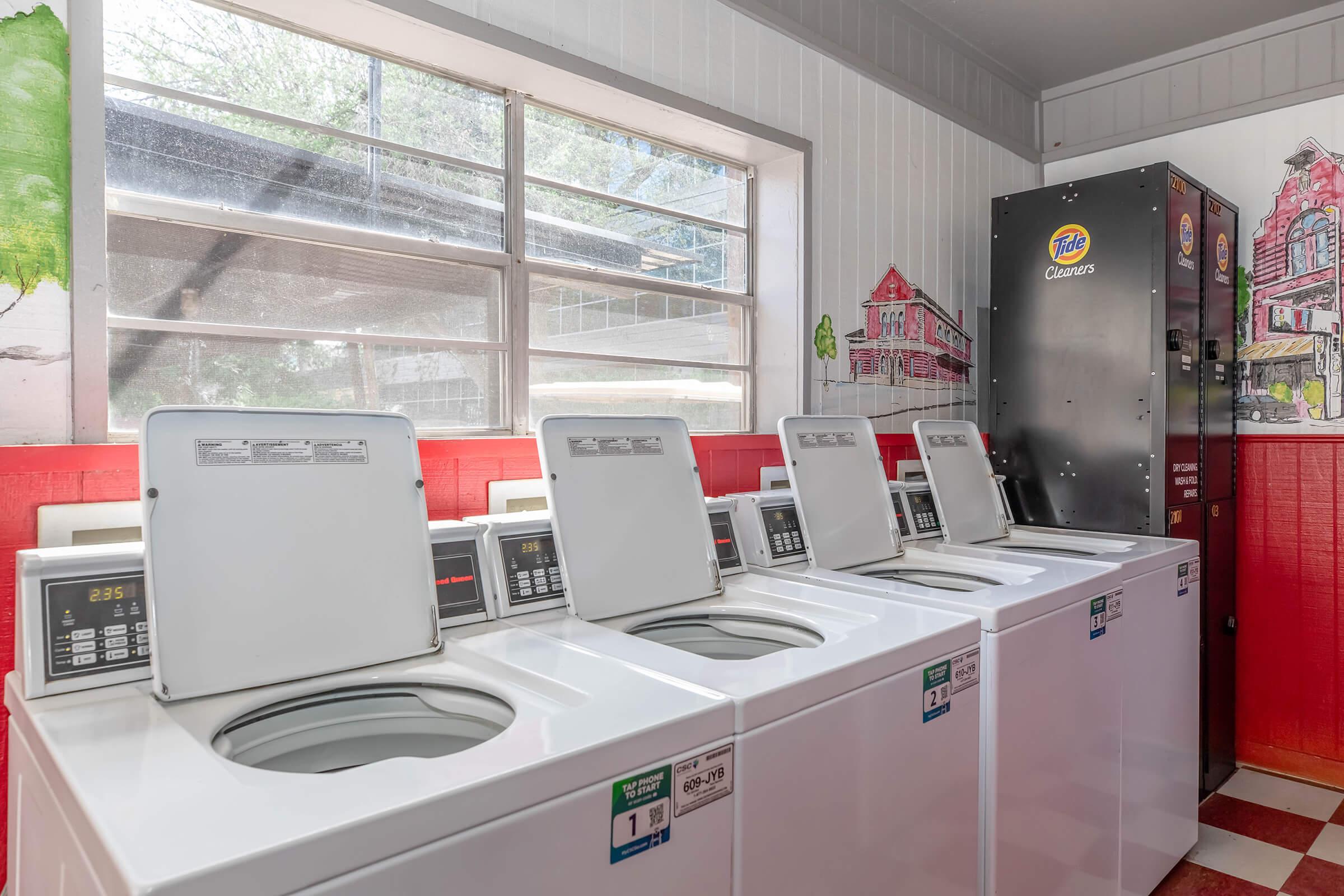 A row of white top-loading washing machines in a brightly colored laundromat, with large windows letting in natural light. A vending machine labeled "Tide Cleaners" is visible in the corner, and the walls are adorned with colorful murals depicting a vibrant street scene.