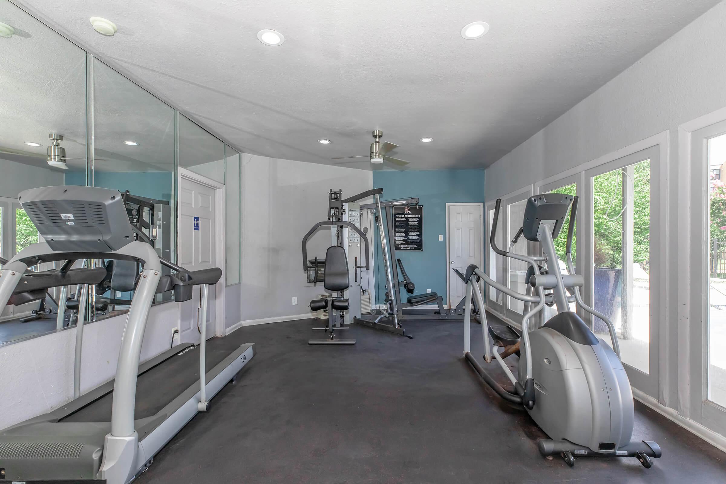 Interior of a fitness room featuring various exercise equipment, including a treadmill, elliptical machine, and strength training station. The space has large mirrors on one wall and windows that provide natural light. The walls are painted light blue, and the flooring is dark.