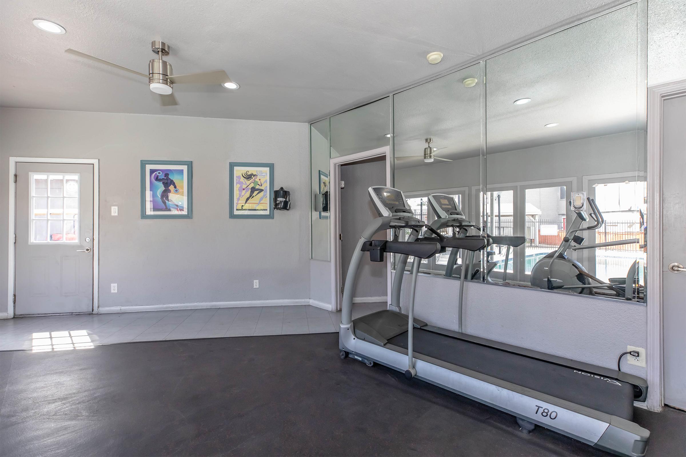 A modern gym area featuring a treadmill, mirrored walls, and colorful framed artwork. A door leads outside, and there are windows allowing natural light in. The floor is a dark shade, contrasting with the light-colored walls. The space appears clean and organized, ideal for workouts.