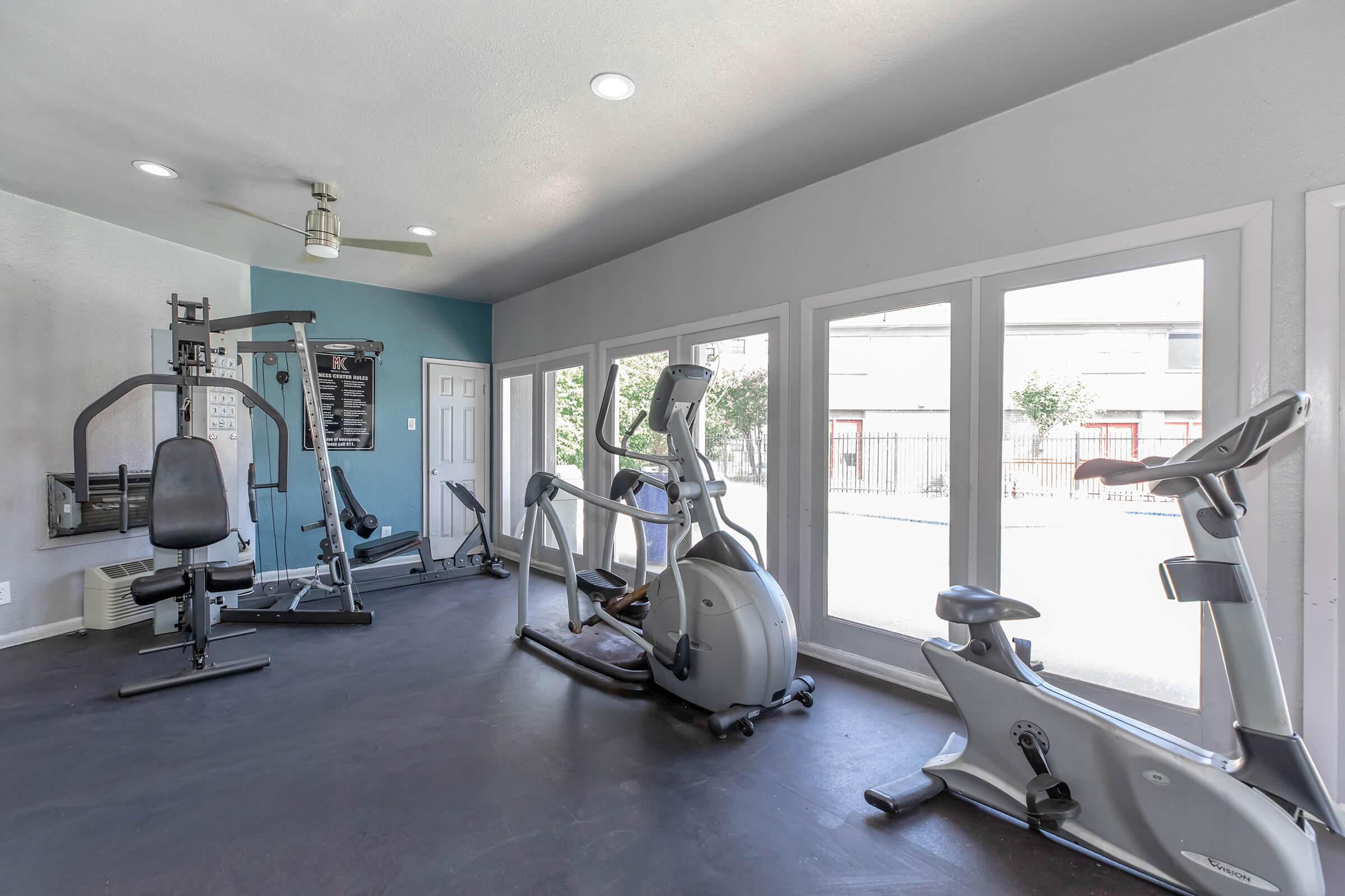 A modern gym interior featuring various exercise equipment, including a stationary bike, elliptical machine, and strength training apparatus. Large windows allow natural light, and a fan is installed on the ceiling. The walls are painted in light colors, creating a bright and inviting workout space.