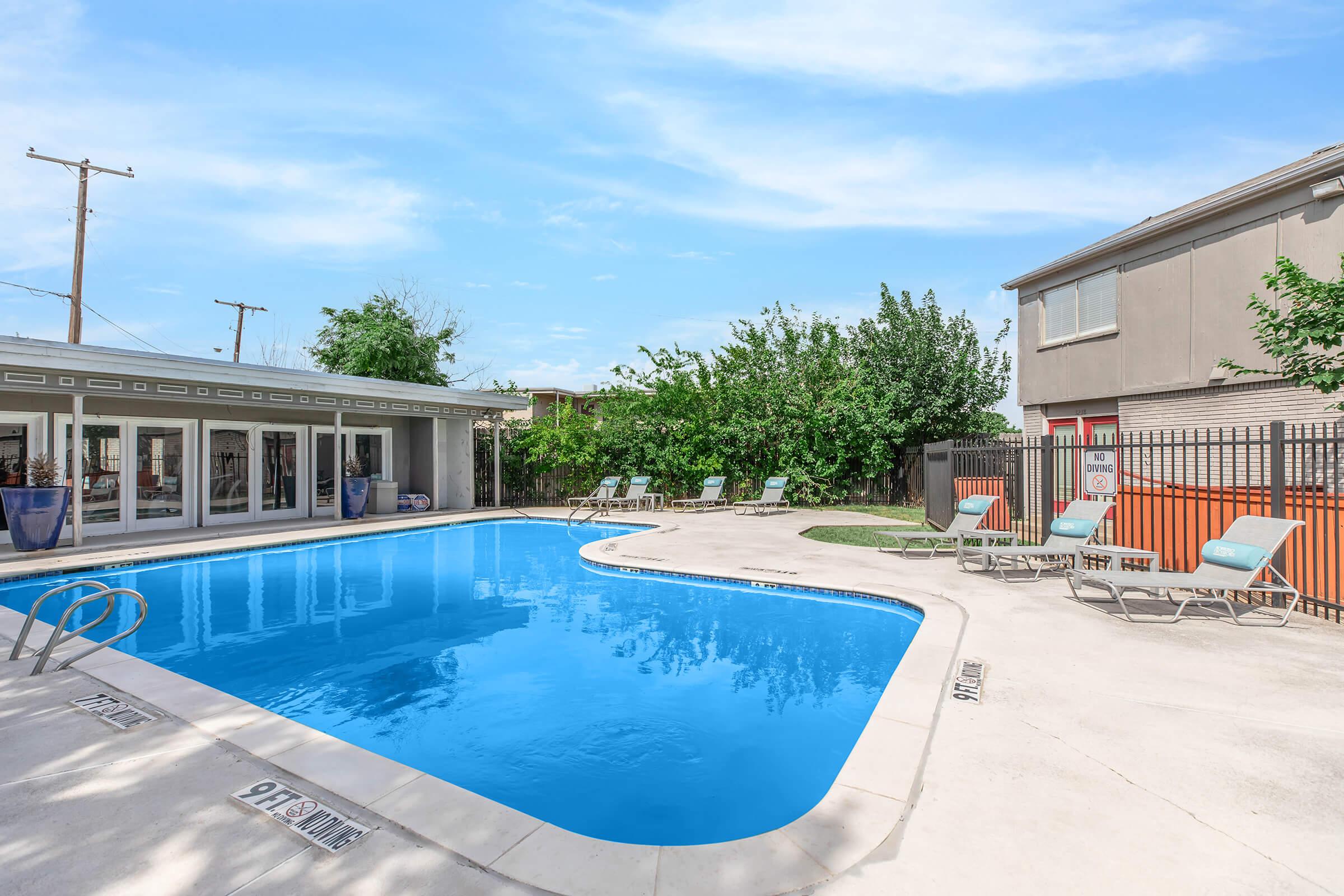 A clear blue swimming pool surrounded by lounge chairs in a well-maintained outdoor area. Lush greenery and a fence are visible in the background, along with a building structure. The sky is bright and sunny, creating a welcoming atmosphere.