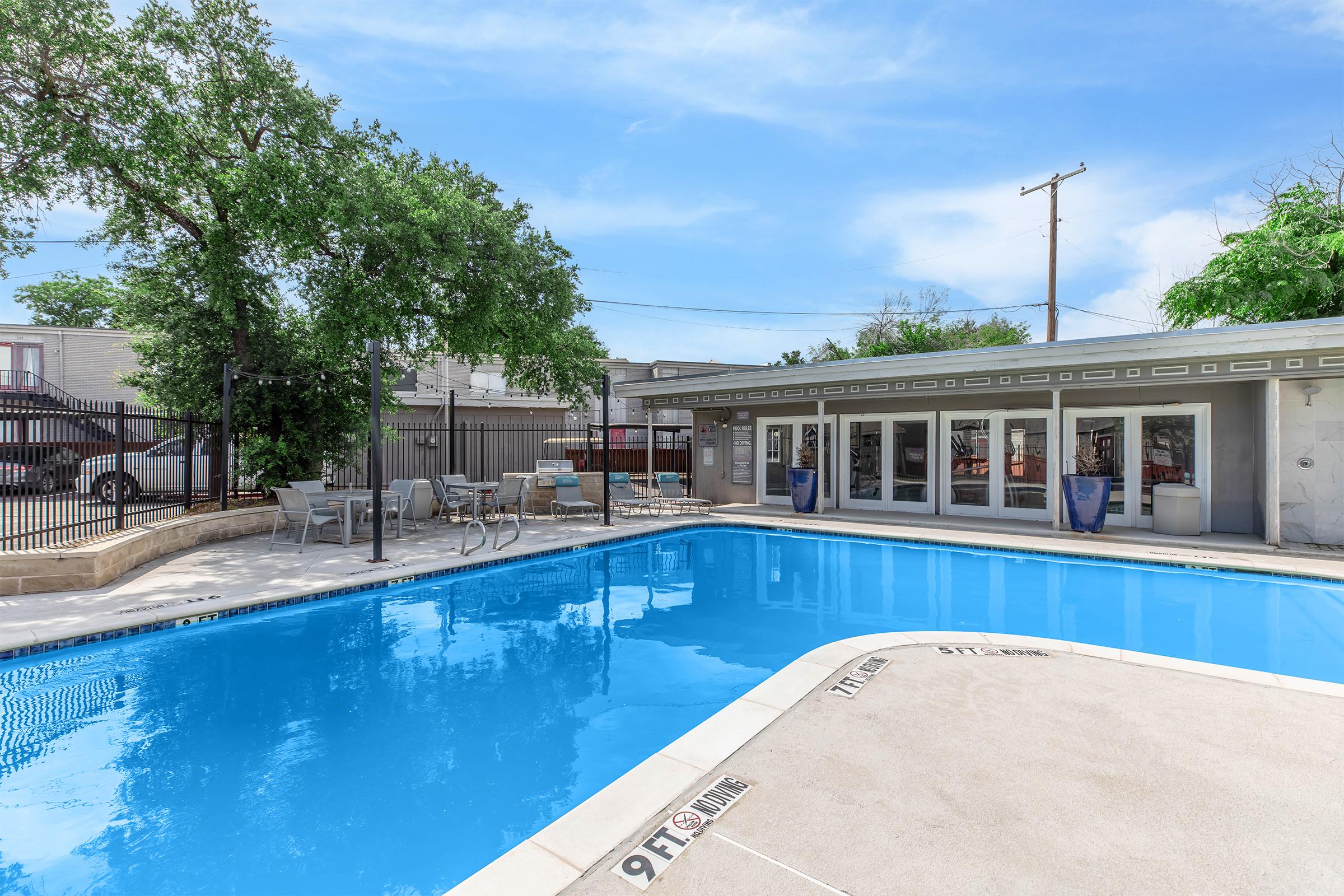 A clear blue swimming pool surrounded by a patio area with lounge chairs. Trees provide shade on one side, and a building with large windows is visible nearby. The pool features marked depth indicators. The sky is partly cloudy, creating a bright and inviting atmosphere.