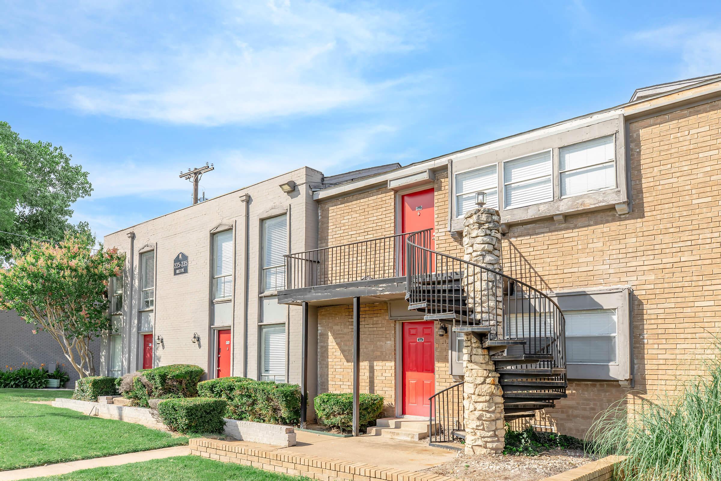 A multi-unit apartment building featuring a two-tone brick facade, red doors, and a spiral staircase. The entrance is flanked by well-maintained shrubs and grass, with a clear blue sky in the background. The building has windows covered with blinds, providing a clean and inviting appearance.
