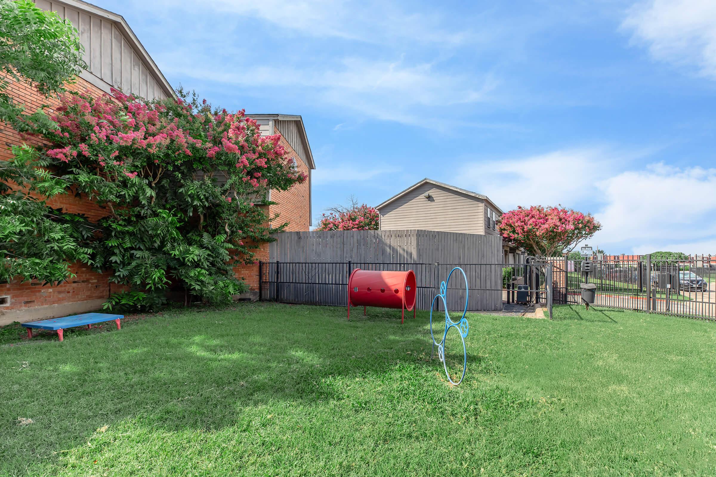 A grassy yard featuring a red play tunnel and a blue bench, surrounded by blooming trees and a wooden fence. Two adjacent buildings are visible in the background under a clear blue sky.