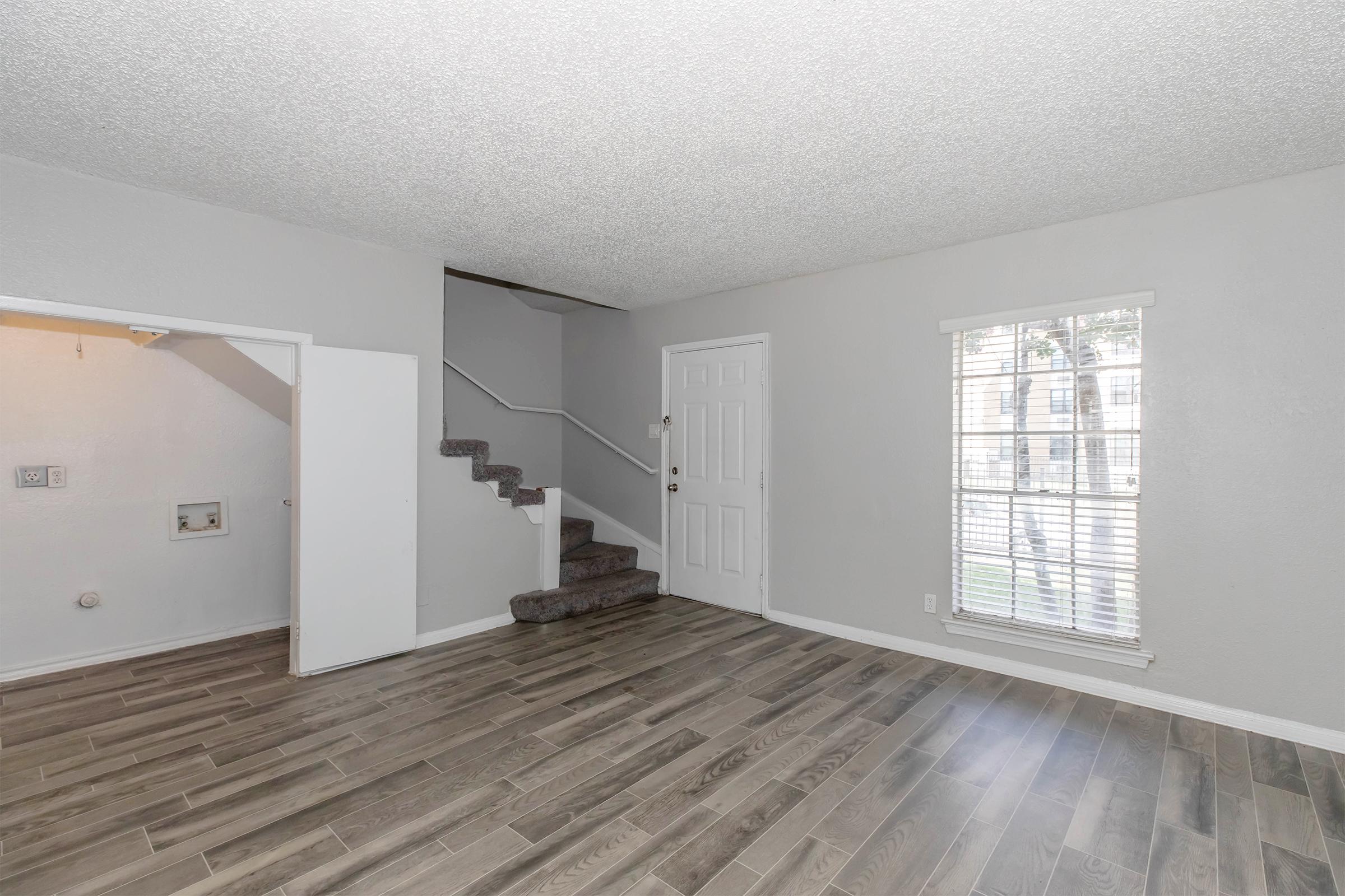 Interior of a living room featuring a staircase, hardwood floors, and a large window with sheer curtains. The walls are painted light gray, and there is a small closet or storage space visible to the left. The overall ambiance is bright and spacious.