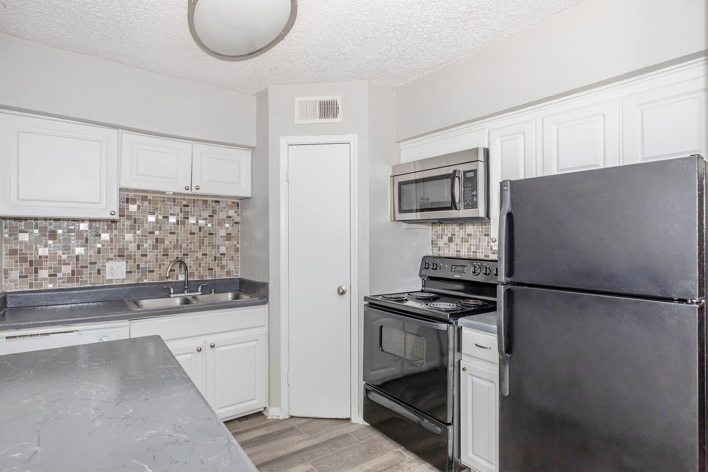 A modern kitchen featuring white kitchen cabinets, a black refrigerator, and a stainless steel microwave. The countertop is gray, and the backsplash consists of mosaic tiles in various colors. A single basin sink is located under a window, and the flooring is a wood-like laminate.