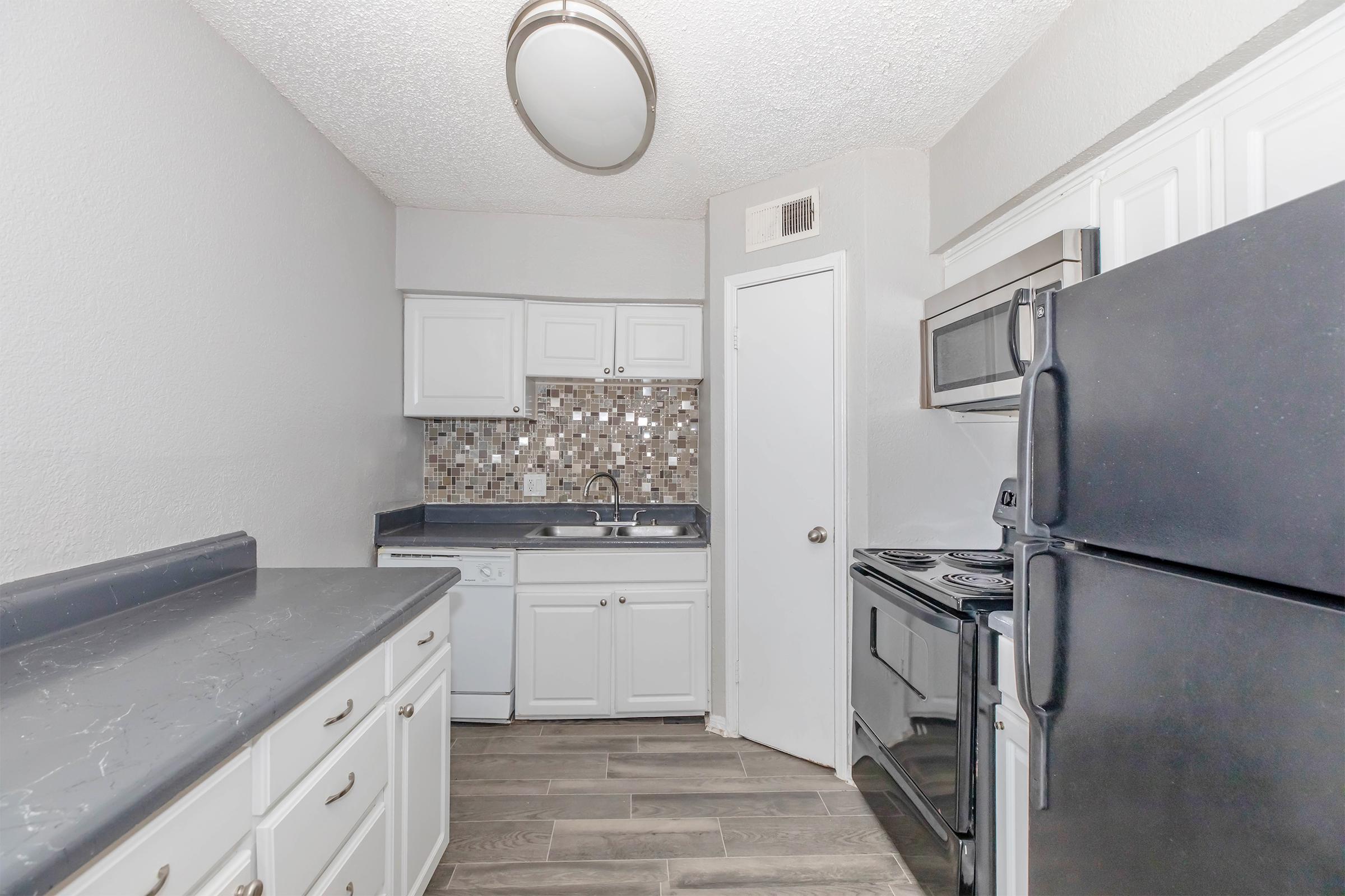 Modern kitchen featuring white cabinetry, dark countertops, a stainless steel microwave, and black appliances. The backsplash consists of mosaic tiles. There are light-colored walls and a ceiling light fixture, creating a bright and open atmosphere.
