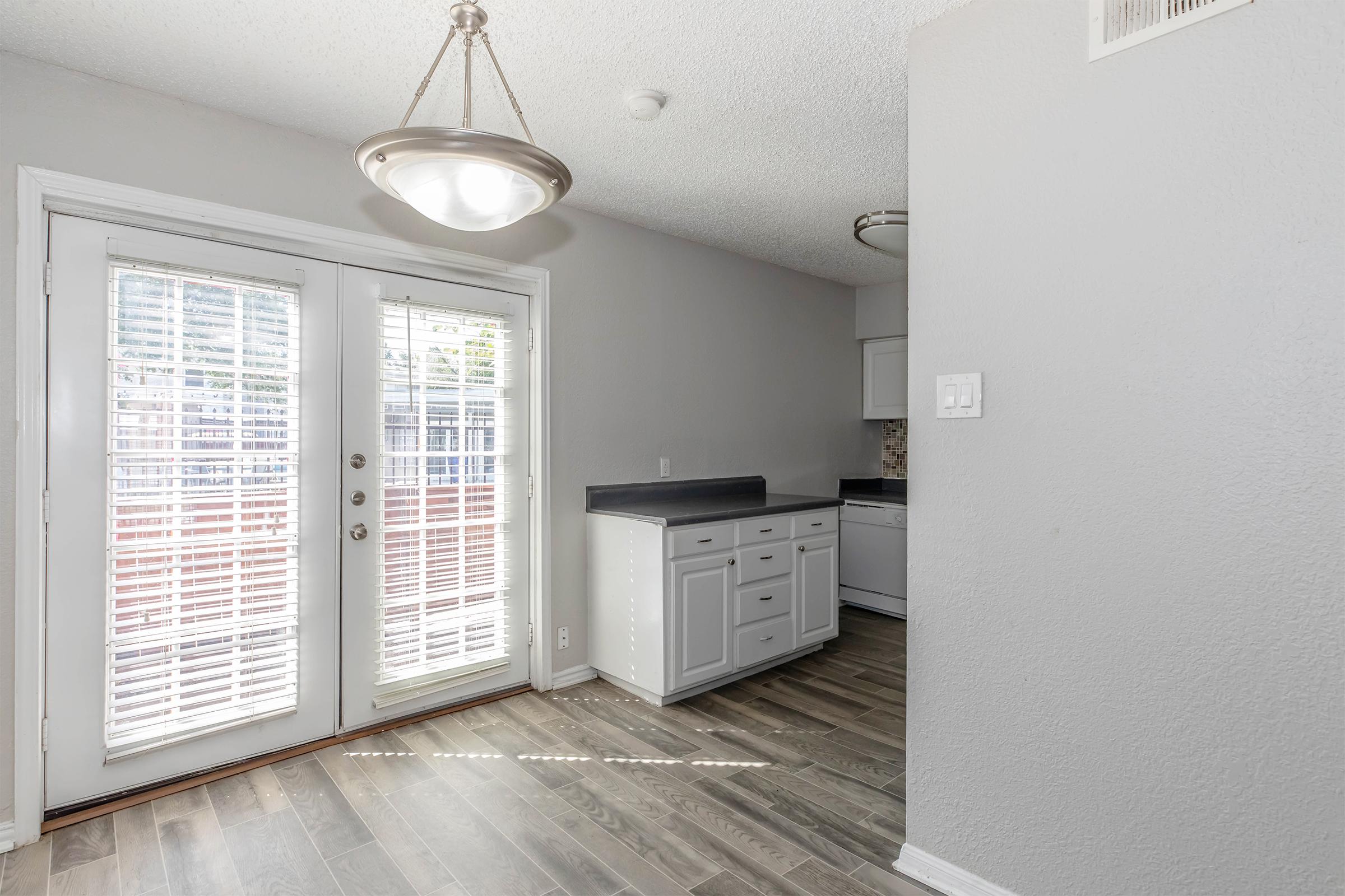 A well-lit dining area featuring a sliding glass door with white shutters, tile floor, and light gray walls. A small kitchen space is visible in the background, with white cabinets and dark countertops. A modern light fixture hangs from the ceiling, enhancing the bright ambiance.
