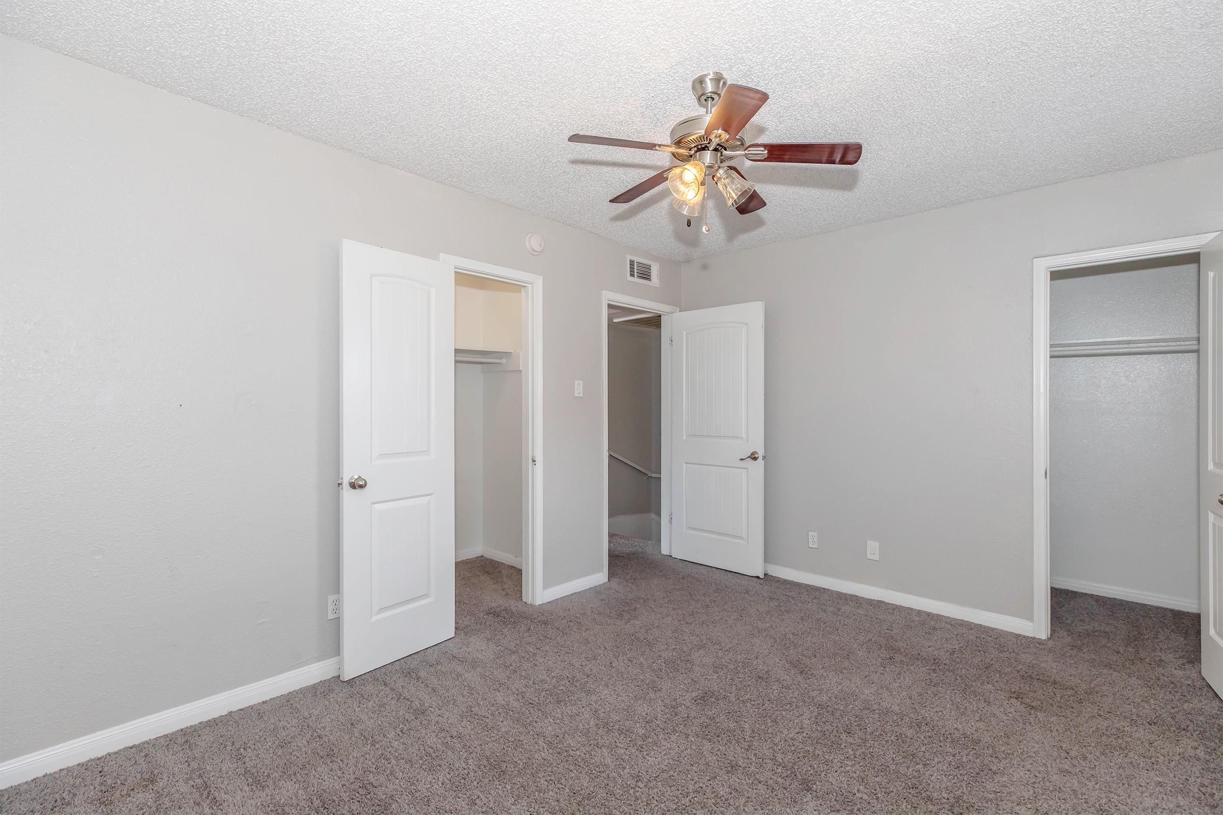 A spacious room featuring light gray walls, a ceiling fan with lights, and plush carpet. Two doors lead to closets, and another door appears to lead outside or to another room. Natural light enters the room through the open doors, creating a bright, inviting atmosphere.