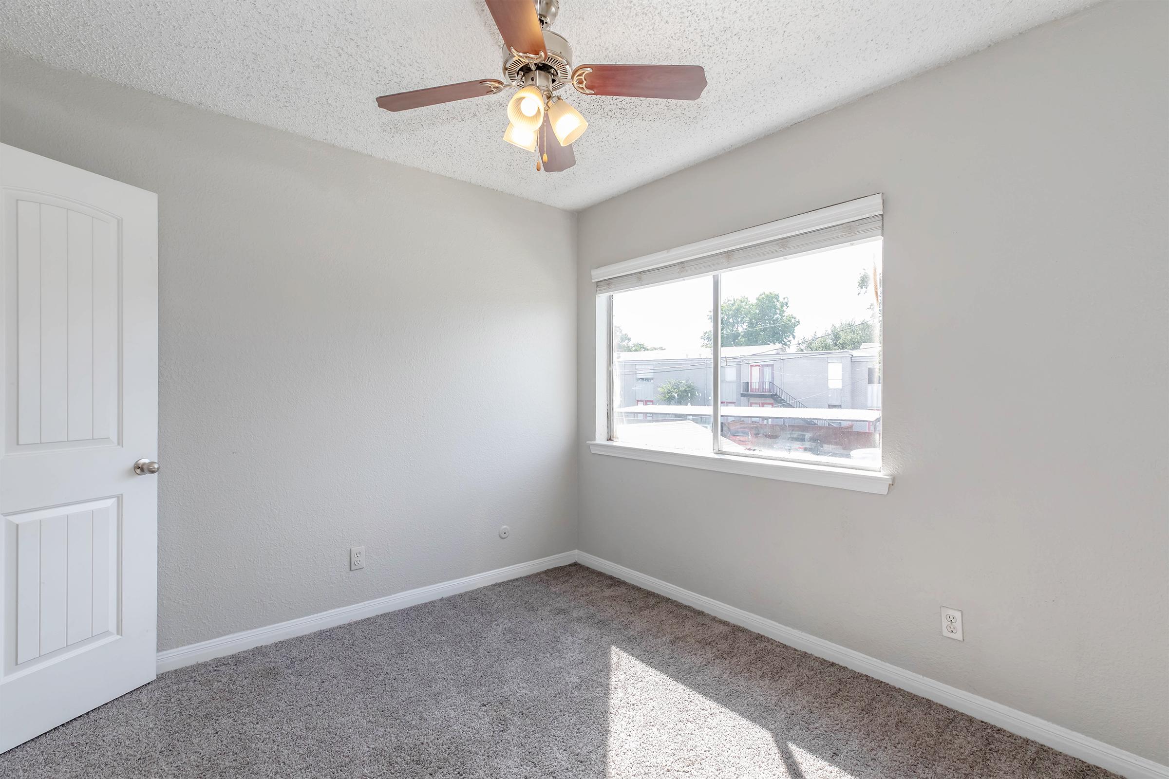 A well-lit room featuring a ceiling fan with three bulbs, a window providing natural light, and soft beige walls. The floor is covered in light gray carpet. A white door is visible on the left side, and there are no furnishings, giving the space a clean, empty look.