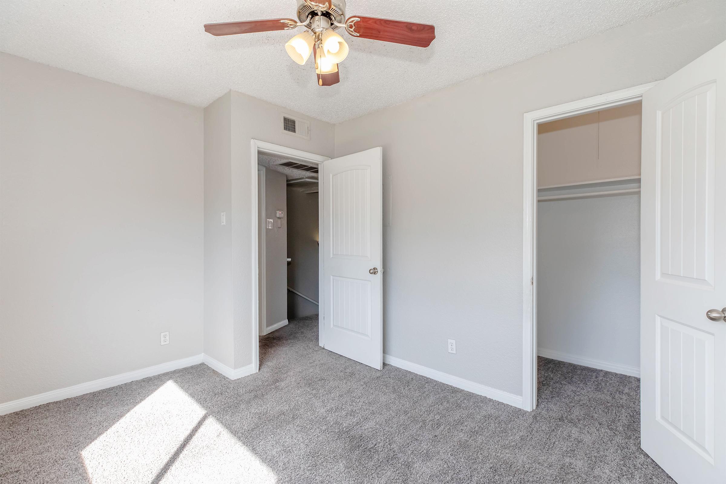A bright and spacious room with light-colored walls and a ceiling fan. The floor is covered with plush carpet. An open door leads to a closet, and another door is visible, indicating access to a hallway or another area of the home. Natural light fills the room, creating a welcoming atmosphere.
