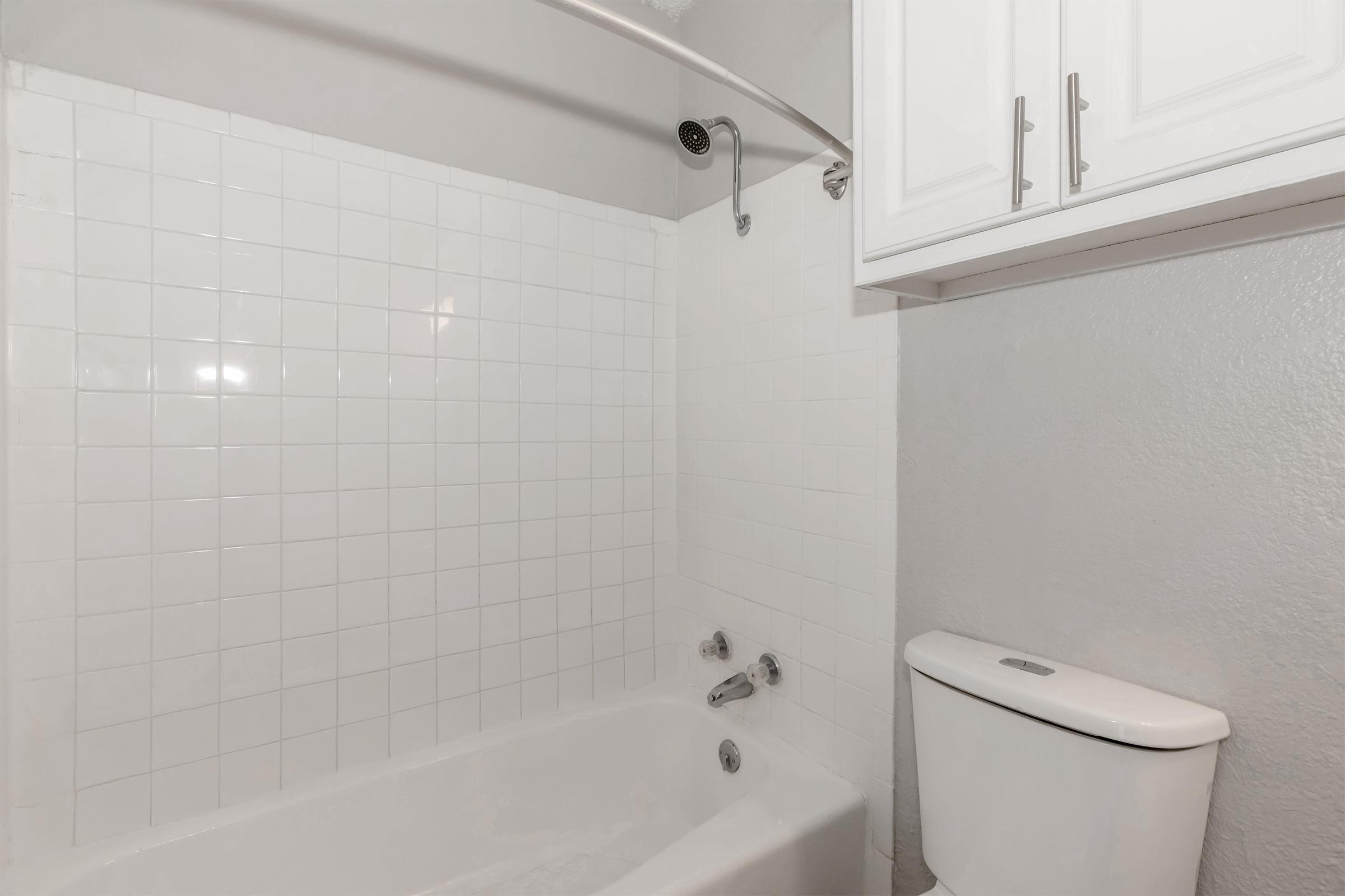 A clean, simple bathroom featuring a white tiled wall, a bathtub with a showerhead, and a white toilet. Above the bathtub, there is a curved shower rod. A small white cabinet is visible on the right side. The overall color scheme is light and neutral.