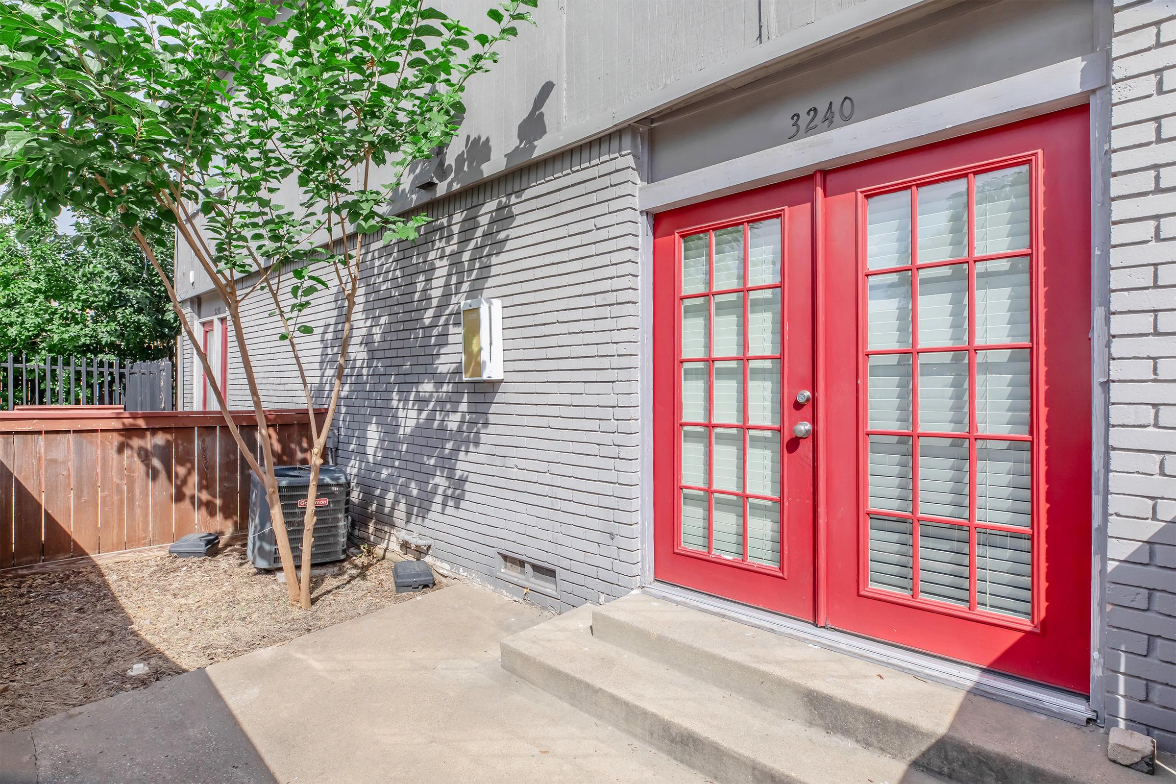 Exterior view of a modern building entrance featuring a pair of red French doors with multiple panes. The area is landscaped with a small tree and gravel, surrounded by a wooden fence. The building's address, 3240, is displayed above the doors. Bright, sunny conditions highlight the entrance.