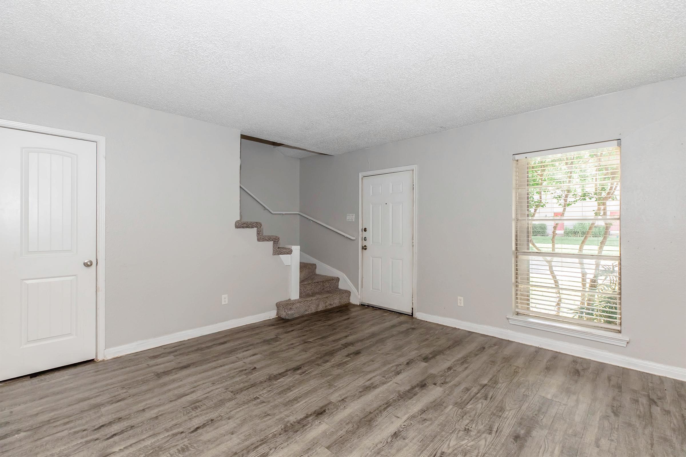 A living room featuring light gray walls and a wood-style floor. There is a staircase leading to the upper level, a closed white door on the left, and a window on the right allowing natural light to enter. The space is minimalist and inviting.