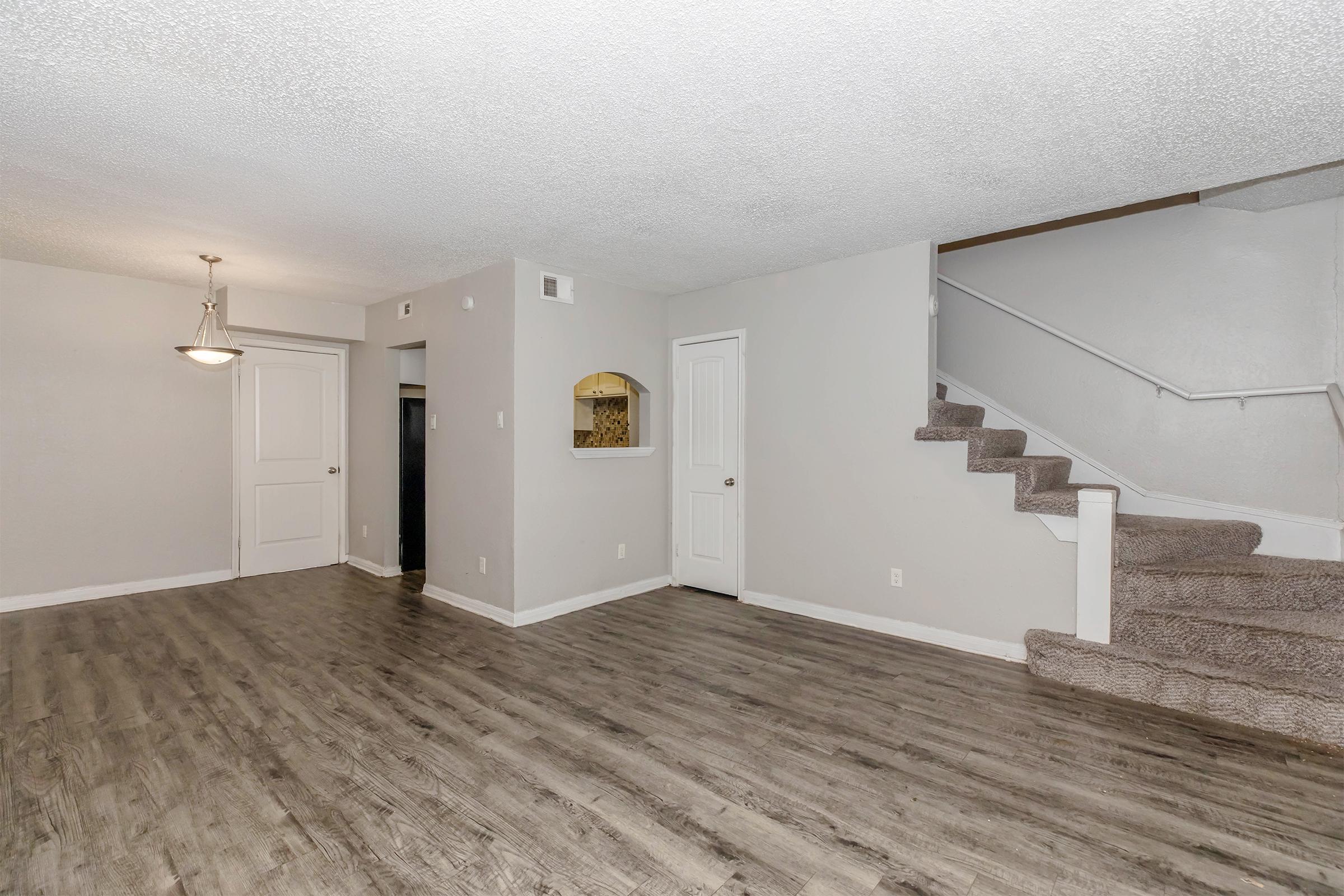A spacious living area featuring gray walls, wooden flooring, and carpeted stairs. To the left, there's a doorway leading to another room, and in the back, a kitchen is visible through an archway. A ceiling light fixture hangs above, and the overall ambiance is modern and inviting.