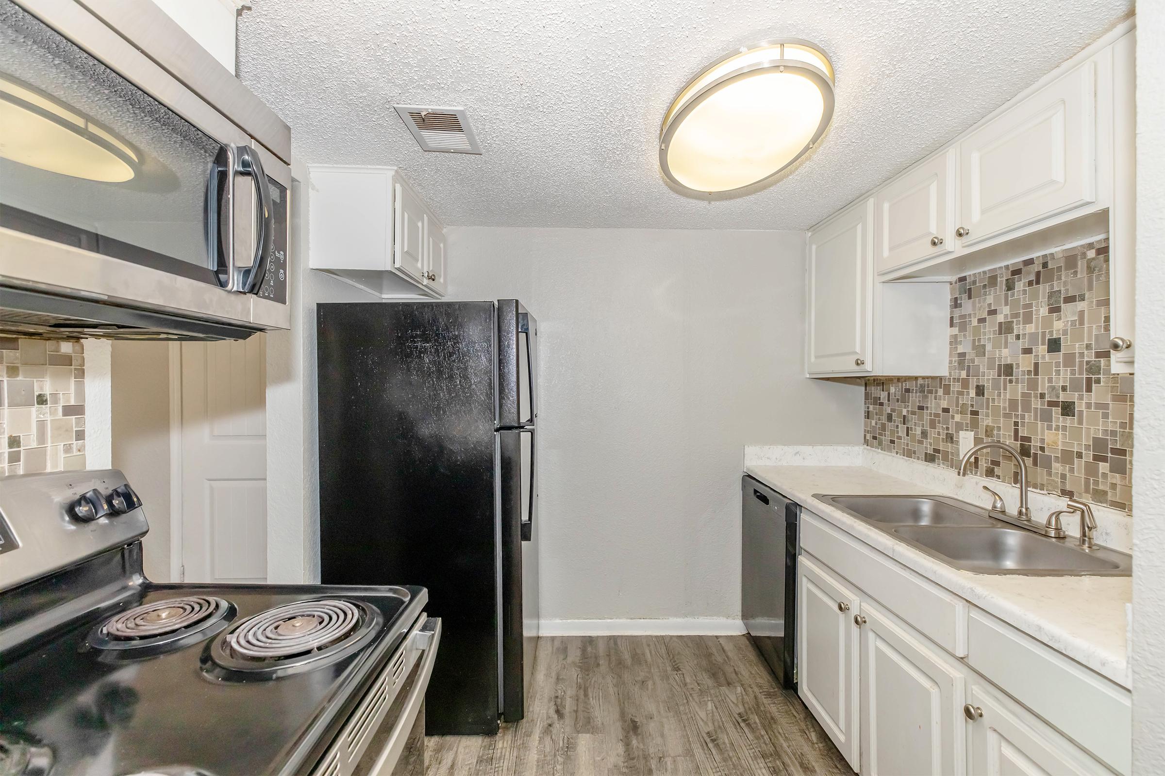 A modern kitchen featuring white cabinetry, a stainless steel stove, an oven, and a black refrigerator. The countertops are light-colored, and there are dual sinks with a modern faucet. The backsplash consists of mosaic tiles in varying shades, complementing the overall design.