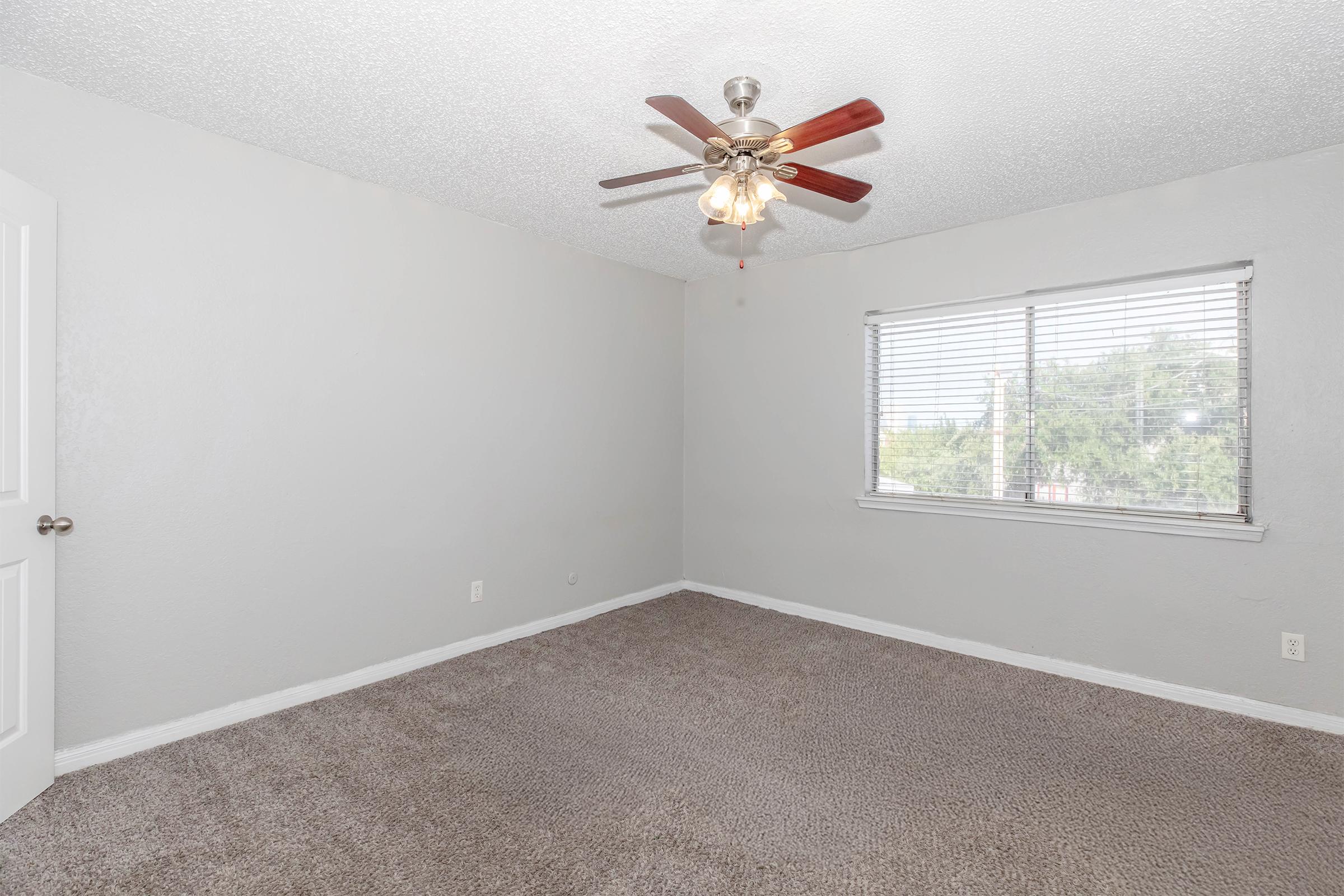 A spacious, empty bedroom with light gray walls and a textured ceiling. The room features a ceiling fan with wooden blades, carpeting in a neutral color, and a window with horizontal blinds allowing natural light in. The overall ambiance is bright and airy, ideal for personal decoration.