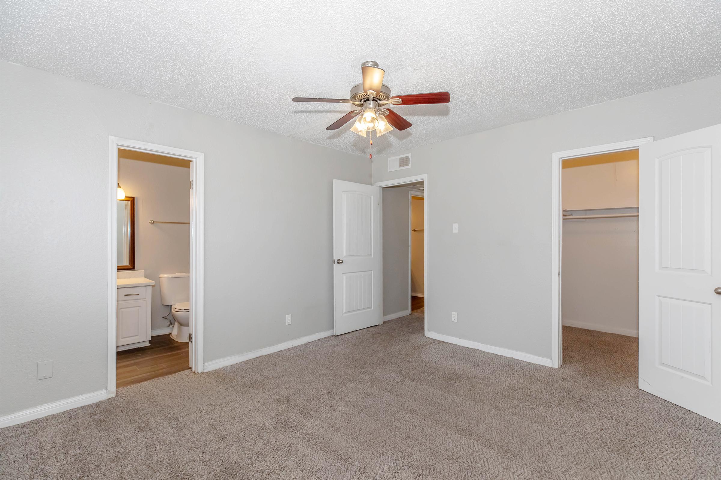 A spacious, neutral-colored room with carpet flooring, featuring a ceiling fan. There are two doors leading to closets and a bathroom, along with a doorframe leading to another area. The walls are light gray and the room is well-lit, creating a cozy atmosphere.