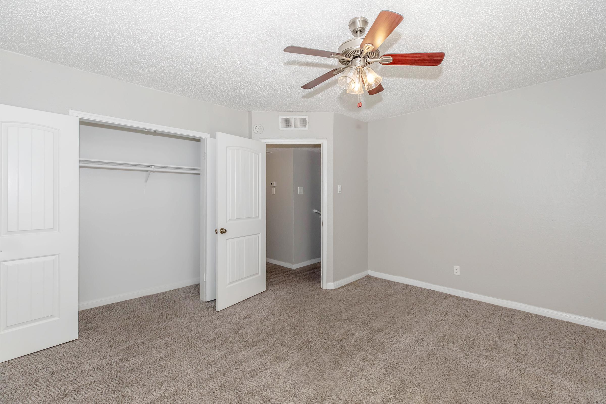 Empty bedroom featuring light gray walls, a ceiling fan with wooden blades, and a plush carpet. There is a closet with double doors and an open doorway to another room. Natural light fills the space, creating a bright and airy atmosphere.